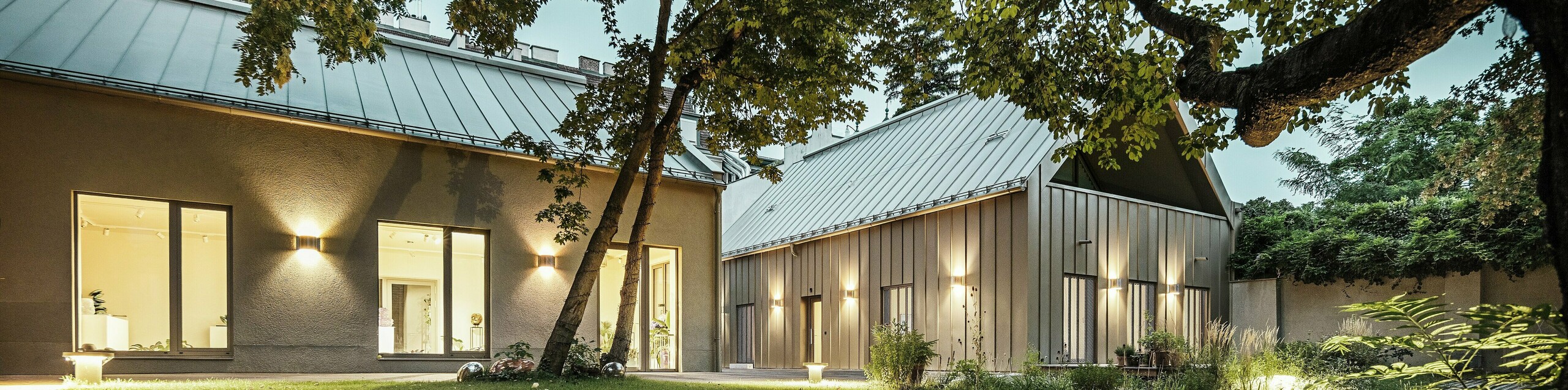 Abendansicht des Studio Comploj in Wien, umgeben von Bäumen und Grünflächen. Die Fassade und das Dach aus PREFALZ-Aluminium in P.10 Prefabronze fügen sich harmonisch in die Umgebung ein.