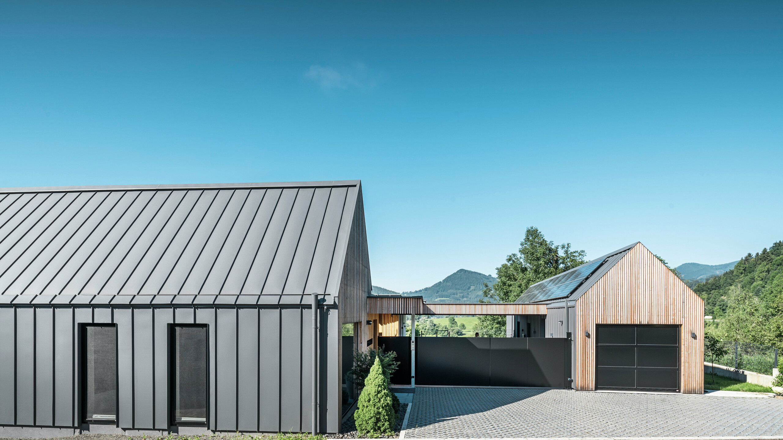 Eingangsbereich eines Einfamilienhauses mit angrenzender Garage in Pogier, Österreich, inmitten einer malerischen Landschaft. Dach und Fassade beider Gebäude sind mit langlebigem PREFALZ in P.10 dunkelgrau verkleidet, was ein modernes Erscheinungsbild erzeugt. Die Garage verfügt über eine harmonisch integrierte Holzverkleidung, die sich nahtlos in die umliegende Landschaft einfügt. Der gepflasterte Hof und die klaren Linien des Entwurfs ergänzen die Ästhetik des Grundstücks, während im Hintergrund die sanften Hügel das Bild einer friedlichen grünen Oase im grünen Herzen Österreichs vervollständigen.