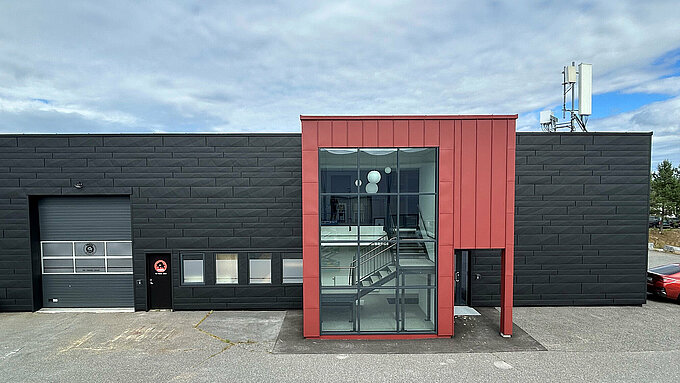 Die Außenansicht des PREFA Academy Gebäudes in Skien, das durch seine moderne Architektur mit einer markanten rot-schwarzen Fassadengestaltung auffällt. Die großen Fenster im oberen Bereich bieten einen Blick auf den Eingangsbereich. Das PREFA-Logo ist gut sichtbar an der Eingangstür angebracht.
