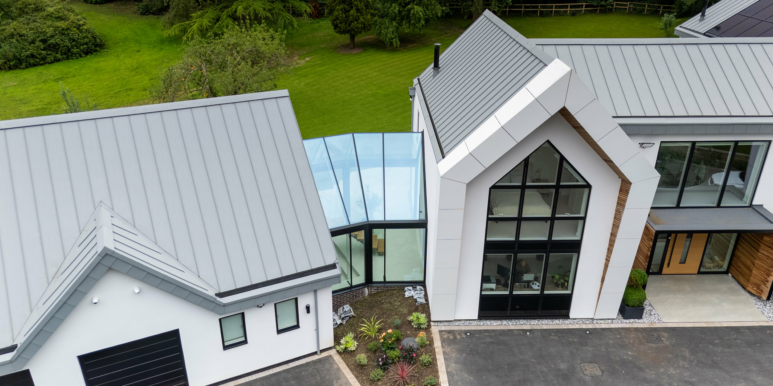 Die Luftaufnahme zeigt eine beeindruckende Ansicht der modernen Villa in Draycott In The Clay, mit ihrem markanten PREFALZ-Dach in Patinagrau. Die symmetrische Stehfalzstruktur des Dachs hebt das Gebäude hervor und fügt sich nahtlos in die Umgebung ein. Besonders auffällig ist die Glasverbindung zwischen den beiden Gebäudeteilen, die den Raum mit Licht durchflutet und das innovative Design betont. Diese Verbindung zeigt eine harmonische Mischung aus modernem Bau und umweltbewusster Architektur, mit Solarpanels, die das Dach ergänzen und die nachhaltige Ausrichtung des Hauses unterstreichen.