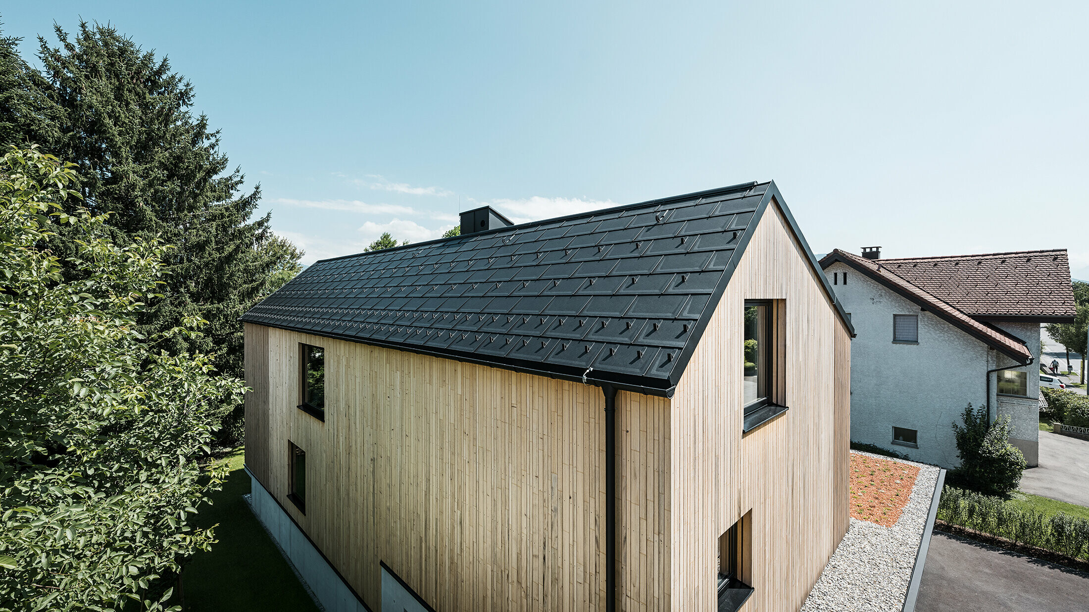 Ein modernes Einfamilienhaus in Lustenau besticht durch die Kombination einer freundlichen Holzfassade mit robusten PREFA Aluminiumprodukten. Das Satteldach wird von der PREFA Dachplatte R.16 mit Schneestoppern sowie der passenden Dachentwässerung in Form von Hängerinne und Ablaufrohr geschmückt. Alle Produkte sind einheitlich im Farbton Schwarz P.10 gehalten und harmonieren perfekt mit der vertikalen Holzfassade.