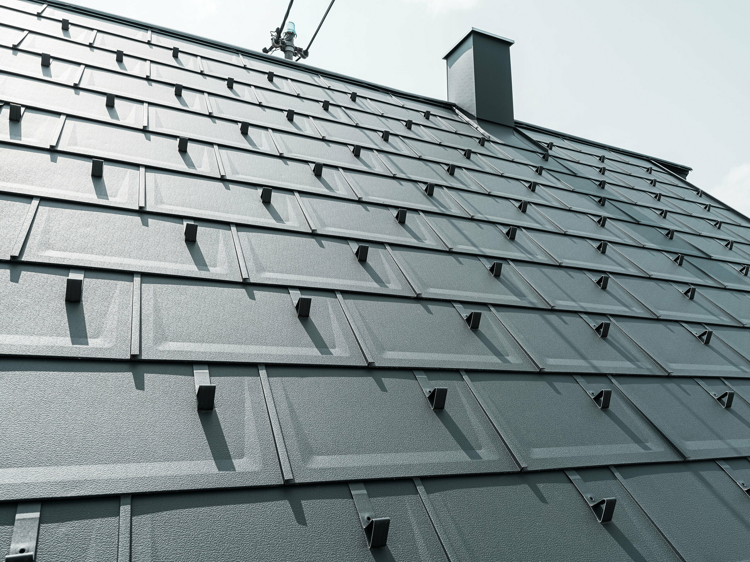 Detaljbilde av PREFA Takplate R.16 i fargen P.10 Antrasitt. På taket ser man PREFA snøfangere og en røykfang.