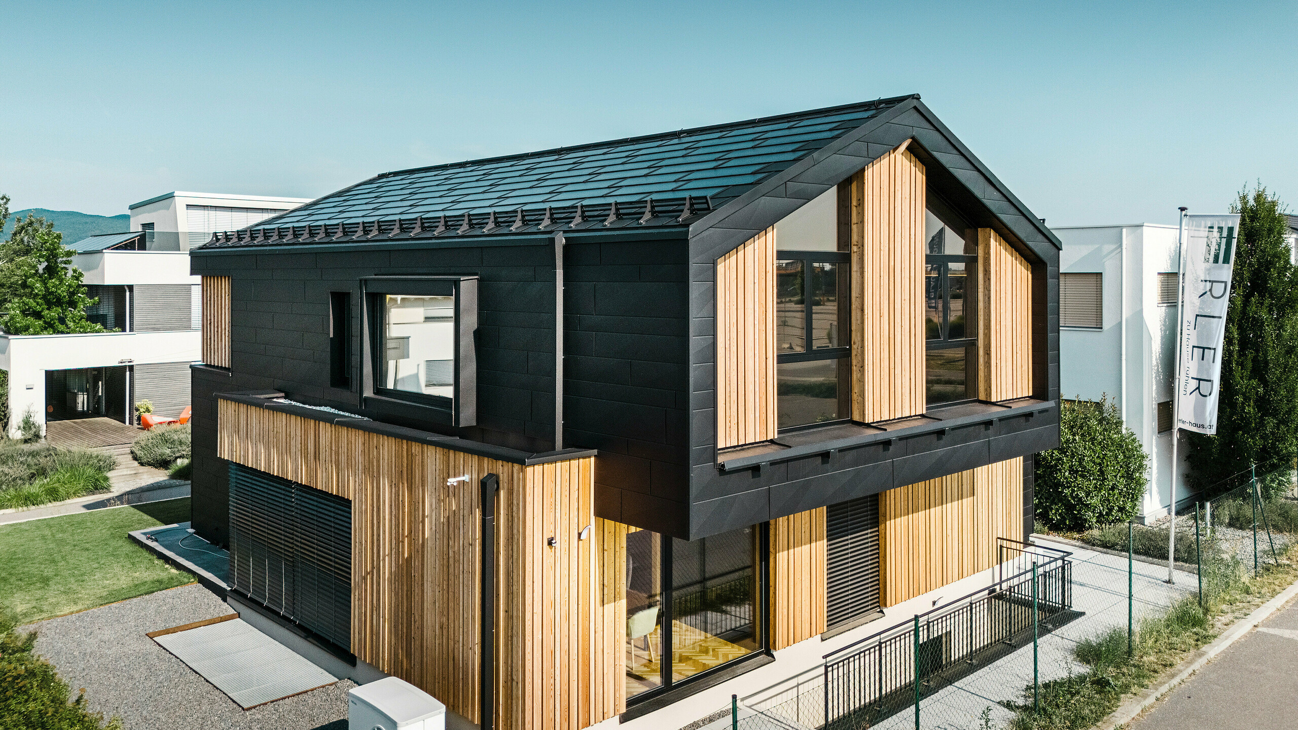 framside av mønsterhuset Autark i prefabrikksenteret Blaue Lagune, Vösendorf (AT). Taket er dekket med svarte PREFA solcelle takplater og takplate R.16 i høy kvalitet P.10-lakkering. Fasade består av en kombinasjon av vertikale treelementer og PREFA Siding.X i P.10 svart, noe som gir bygningen et moderne og bærekraftig preg. På høyre side av bildet vaier flagget til Erler Bau GmbH.