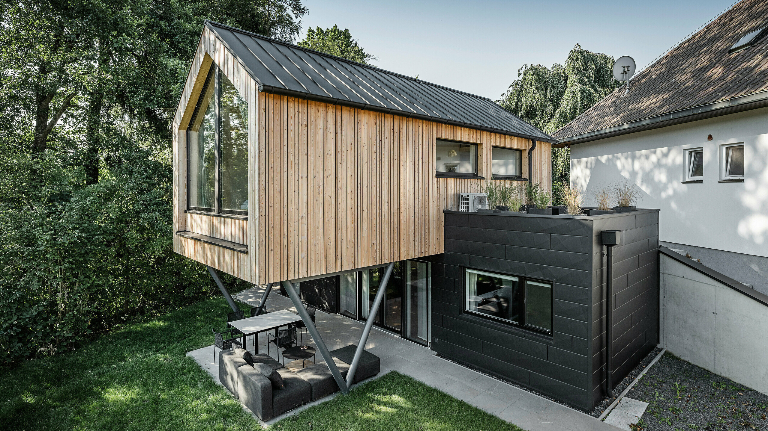 Das 'Woodstock Chalet 5' beeindruckt mit einer eindrucksvollen Kombination aus natürlichen Holzoberflächen und den modernen PREFA Siding.X Fassadenelementen in Anthrazit. Die überhängende Holzstruktur des Obergeschosses ragt markant über die dunklen Fassadenelemente, die das Erdgeschoss verkleiden, und erzeugt so ein Spiel aus Licht und Schatten. Die klaren Linien und die robuste Konstruktion der Siding.X Elemente ergänzen das innovative Design des Gebäudes und bieten einen spannenden visuellen Kontrast zur umgebenden Vegetation und dem traditionellen Gebäude im Hintergrund. Am Dach des Chalets wurde das PREFALZ Dachsystem in Form einer gleichmäßigen Stehfalzdeckung verlegt. Am rechten Rand des Erdgeschosses ist das PREFA Dachentwässerungssystem zu erkennen, das analog zur Aluminiumfassade in Anthrazit gehalten ist. Für die Dachentwässerung wurden der Wasserfangkasten gefolgt vom Ablaufrohr montiert.