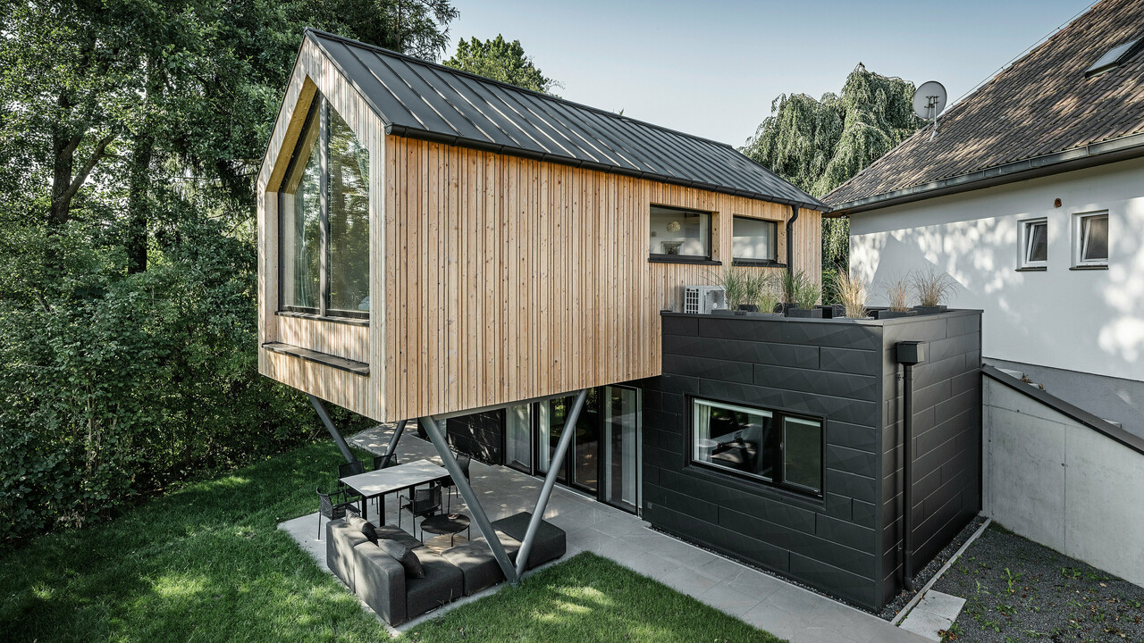 Das 'Woodstock Chalet 5' beeindruckt mit einer eindrucksvollen Kombination aus natürlichen Holzoberflächen und den modernen PREFA Siding.X Fassadenelementen in Anthrazit. Die überhängende Holzstruktur des Obergeschosses ragt markant über die dunklen Fassadenelemente, die das Erdgeschoss verkleiden, und erzeugt so ein Spiel aus Licht und Schatten. Die klaren Linien und die robuste Konstruktion der Siding.X Elemente ergänzen das innovative Design des Gebäudes und bieten einen spannenden visuellen Kontrast zur umgebenden Vegetation und dem traditionellen Gebäude im Hintergrund. Am Dach des Chalets wurde das PREFALZ Dachsystem in Form einer gleichmäßigen Stehfalzdeckung verlegt. Am rechten Rand des Erdgeschosses ist das PREFA Dachentwässerungssystem zu erkennen, das analog zur Aluminiumfassade in Anthrazit gehalten ist. Für die Dachentwässerung wurden der Wasserfangkasten gefolgt vom Ablaufrohr montiert.