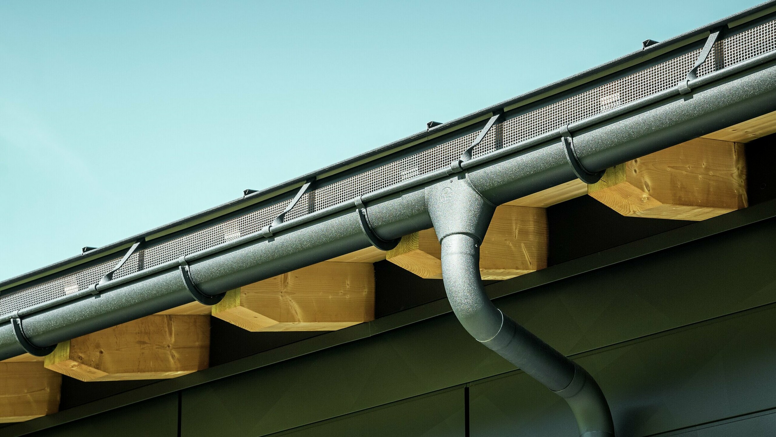 Nærbilde av fasaden på en moderne enebolig i Ascharina, Sveits. Bygningens sider er kledd med de markante PREFA Siding.X fasadepaneler i P.10 svart. PREFA takdrenering med takrenne og nedløpsrør er fargematchende.