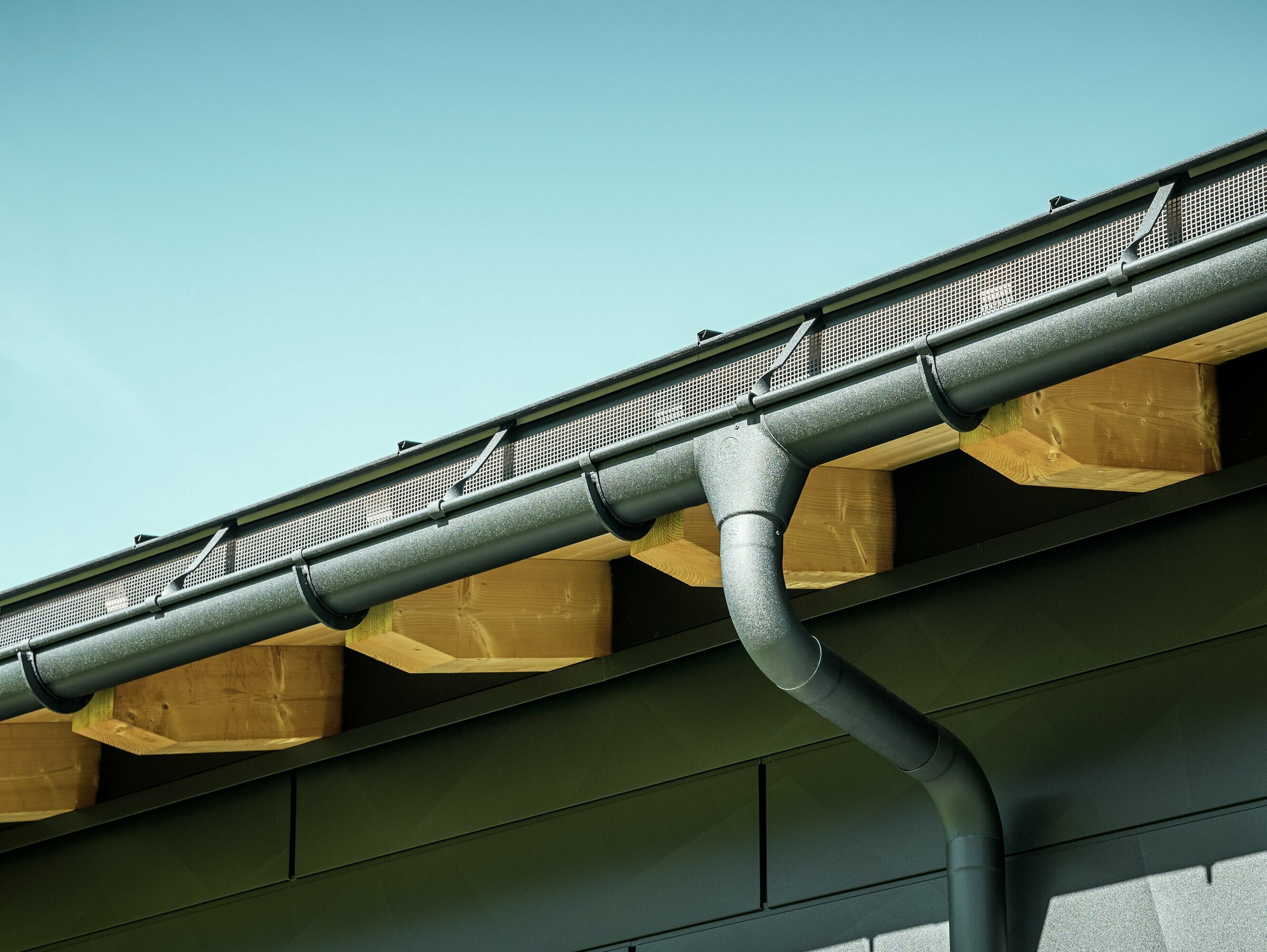 Nærbilde av fasaden på en moderne enebolig i Ascharina, Sveits. Bygningens sider er kledd med de markante PREFA Siding.X fasadepaneler i P.10 svart. PREFA takdrenering med takrenne og nedløpsrør er fargematchende.