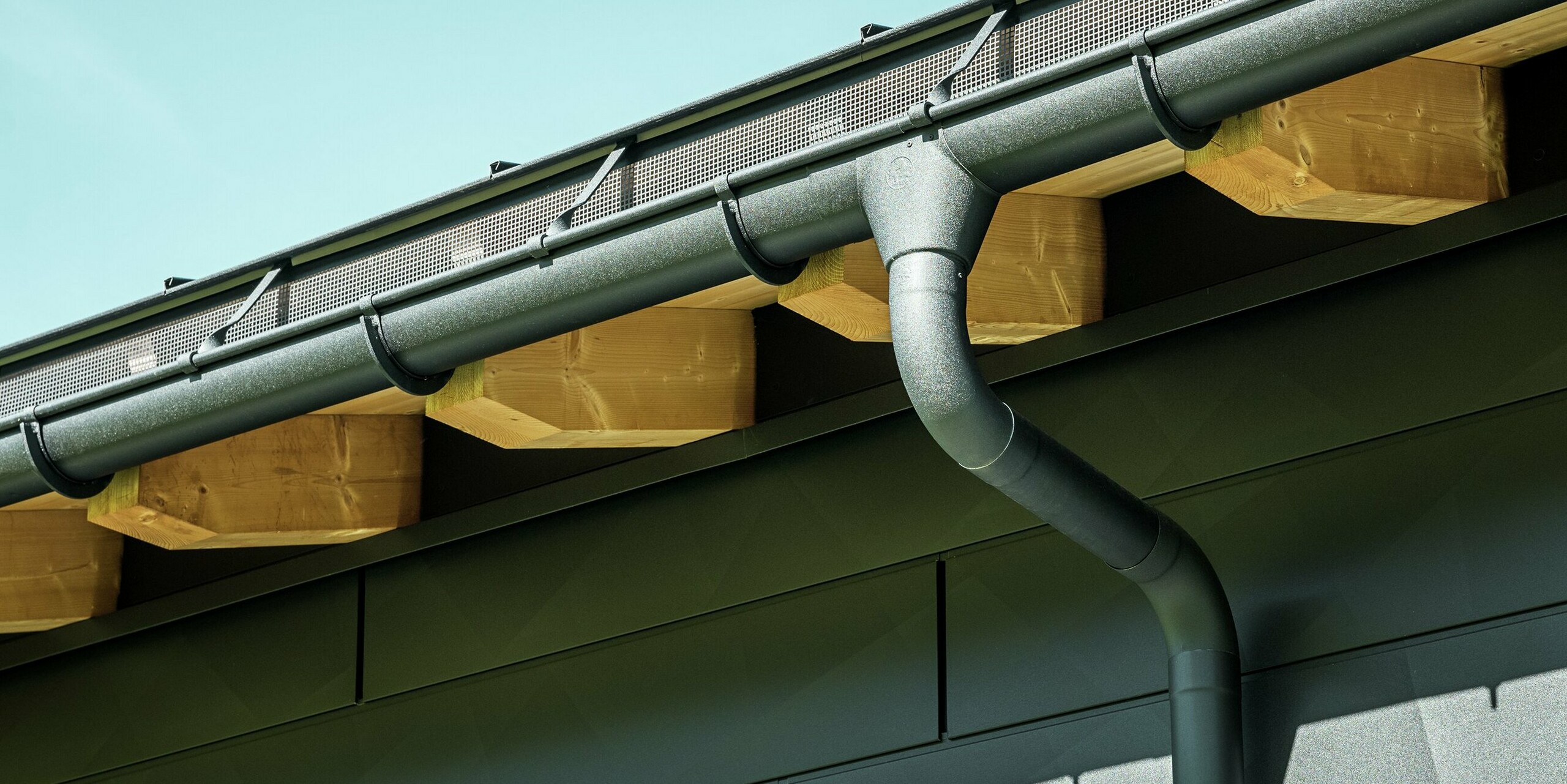 Nærbilde av fasaden på en moderne enebolig i Ascharina, Sveits. Bygningens sider er kledd med de markante PREFA Siding.X fasadepaneler i P.10 svart. PREFA takdrenering med takrenne og nedløpsrør er fargematchende.