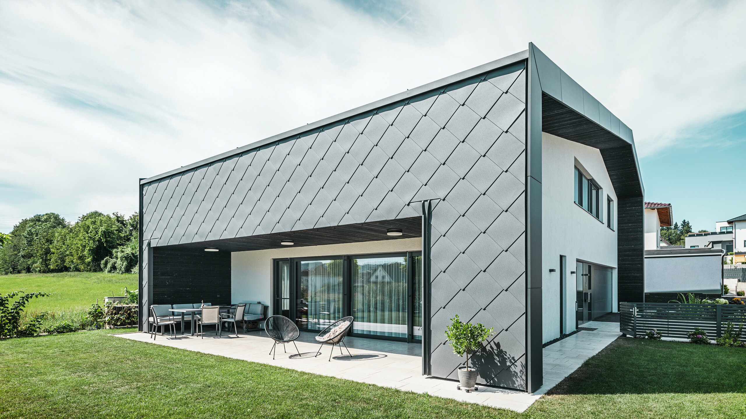 Velstelt hageområde til en enebolig i Aschach. Bygningen er stilfullt kledd med mørk grå PREFA aluminium veggrombe 44 × 44. Den overbygde terrassen gir et koselig sitteplass med utsikt over den frodige grønne plenen. Den elegante aluminiumsbekledningen i kombinasjon med hvite pussflater gir en moderne og værbestandig arkitektur. Store vindusflater slipper mye dagslys inn i rommene og skaper en åpen, lys atmosfære.