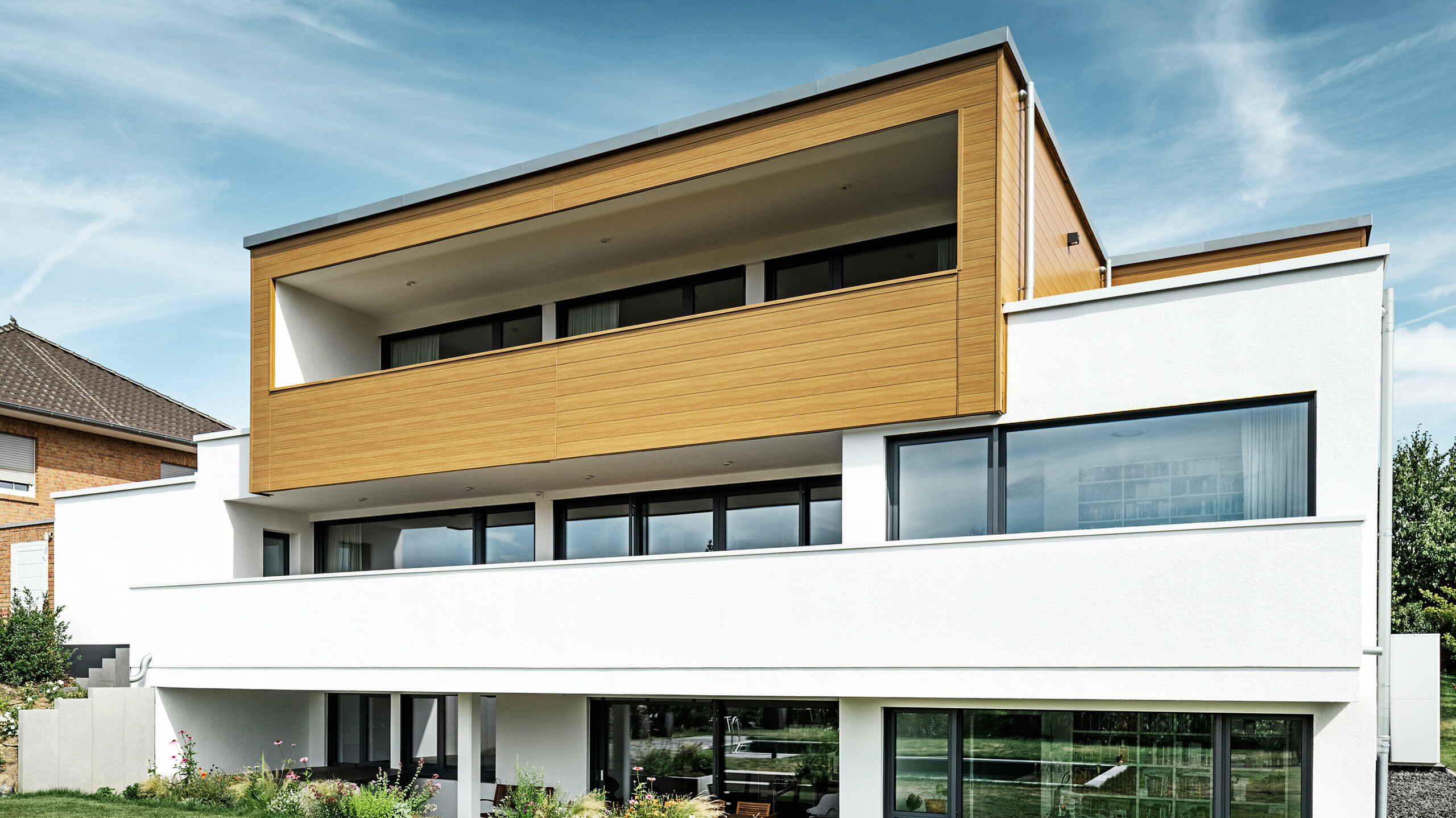 Baksiden av en moderne enebolig i Usingen, Tyskland. Den grønne plenen, bassenget og et ungt hagetre skimtes nederst i bildet. Det iøynefallende huset har en hvit bygningsbase bestående av en pusset fasade, en aluminiumsfasade i øverste etasje laget av PREFA Sidings i natureik og store vindusflater med antrasittfarget ramme. På avstand kan man se nabohuset med teglsteinsfasade i venstre kant av bildet.