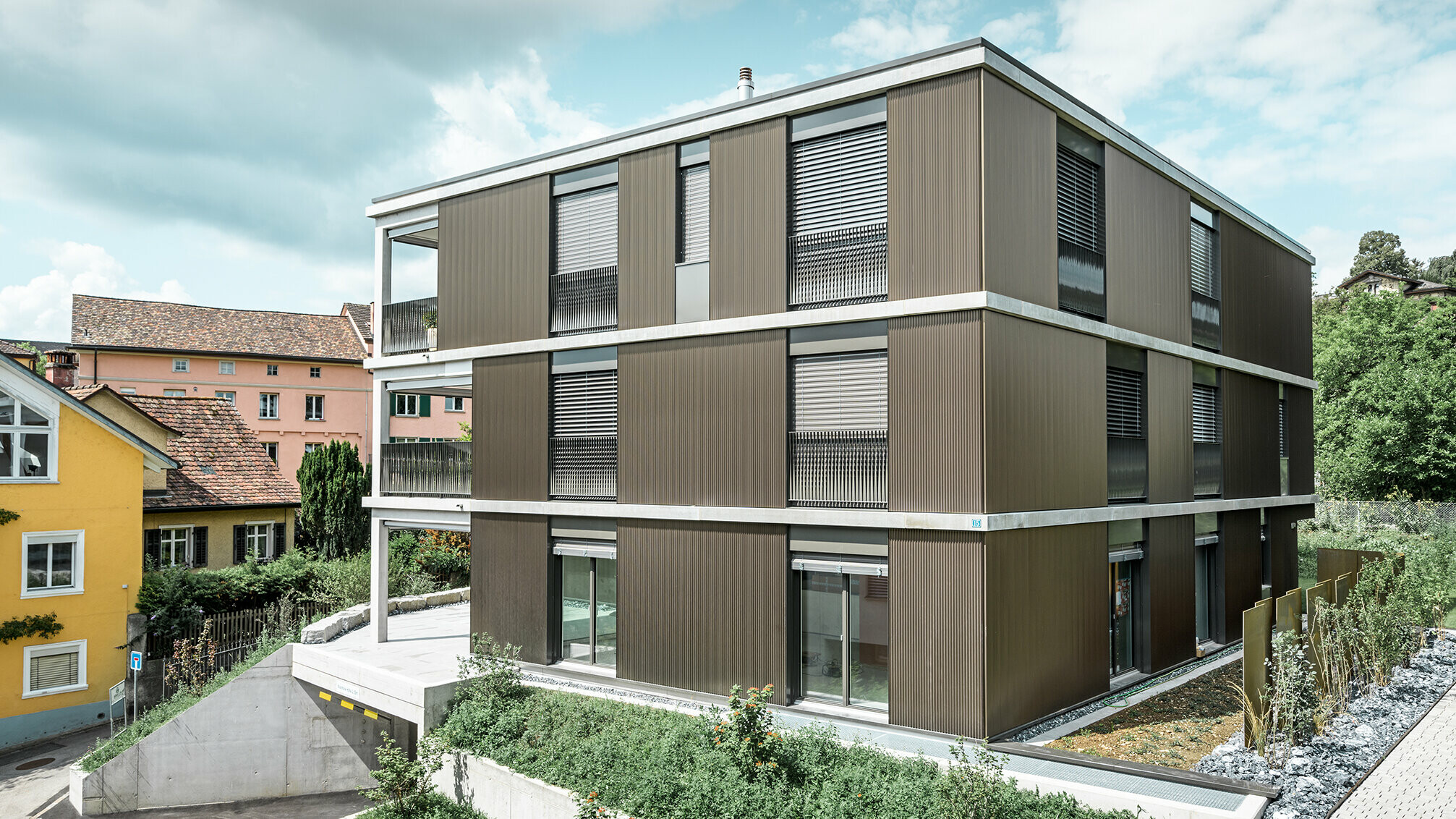 Den markante aluminiumsfasaden på denne flermannsboligen i Schaffhausen er utstyrt med det elegante PREFA-sikksakkprofilet i fargen Colinal. Den vertikale plasseringen av de ekstruderte aluminiumsprofilene gjør at bygningen virker mer kompakt, og fasadestrukturen får en harmonisk balanse. Det elegante designet passer smakfullt inn i omgivelsene og gir bygningen en moderne eleganse som både er visuelt tiltalende og funksjonell. Perspektivet viser flermannsboligen fra framsideen med garasjeinnkjørselen. I bakgrunnen ser man trær, andre bygninger og et kirketårn.