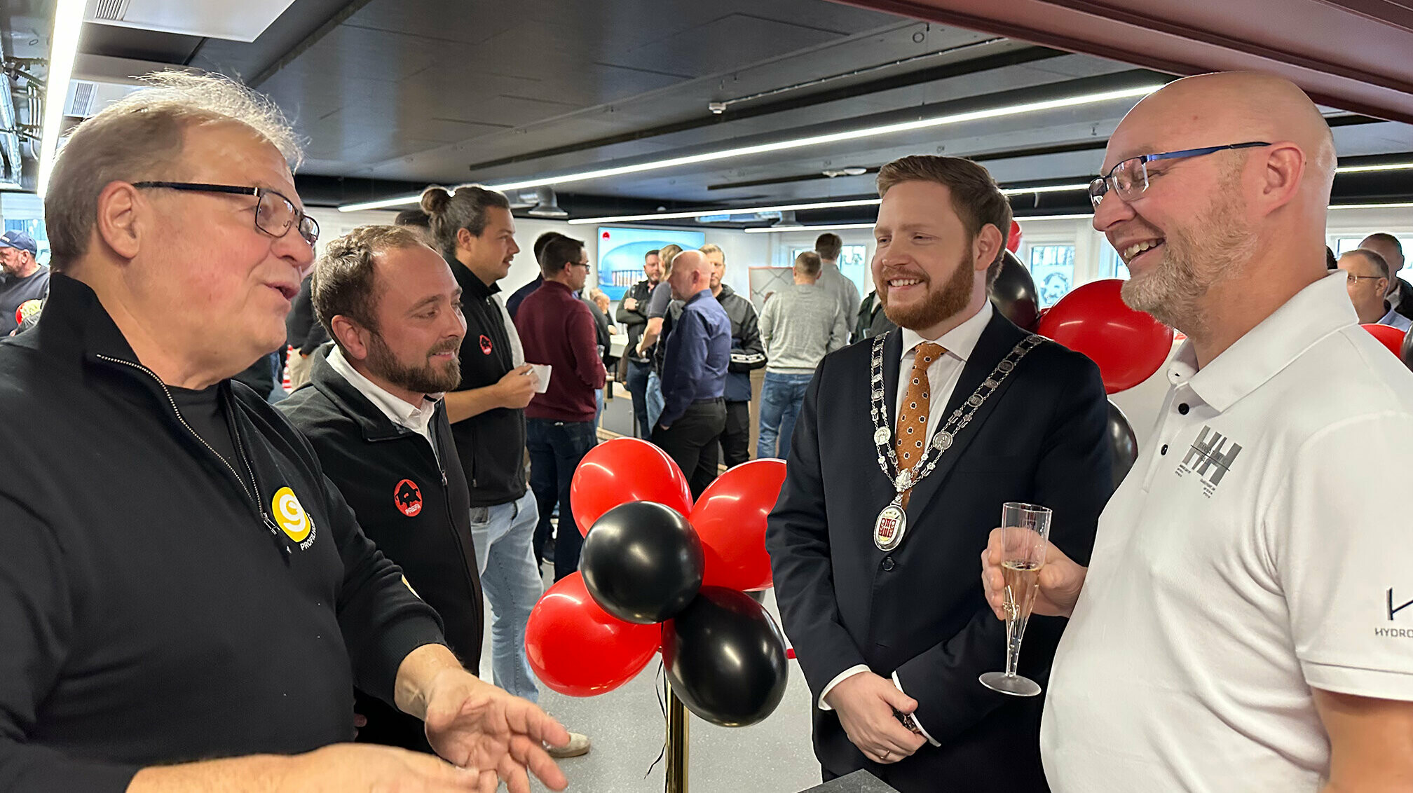 Der Bürgermeister von Skien im Gespräch mit Mitarbeitern und Partnern von PREFA während der Eröffnung der PREFA Academy in Skien. Im Hintergrund sind weitere Gäste der Veranstaltung zu sehen, die in einer festlichen Atmosphäre zusammenkommen. Passend zum PREFA Logo wurden rote und schwarze Luftballons aufgehangen