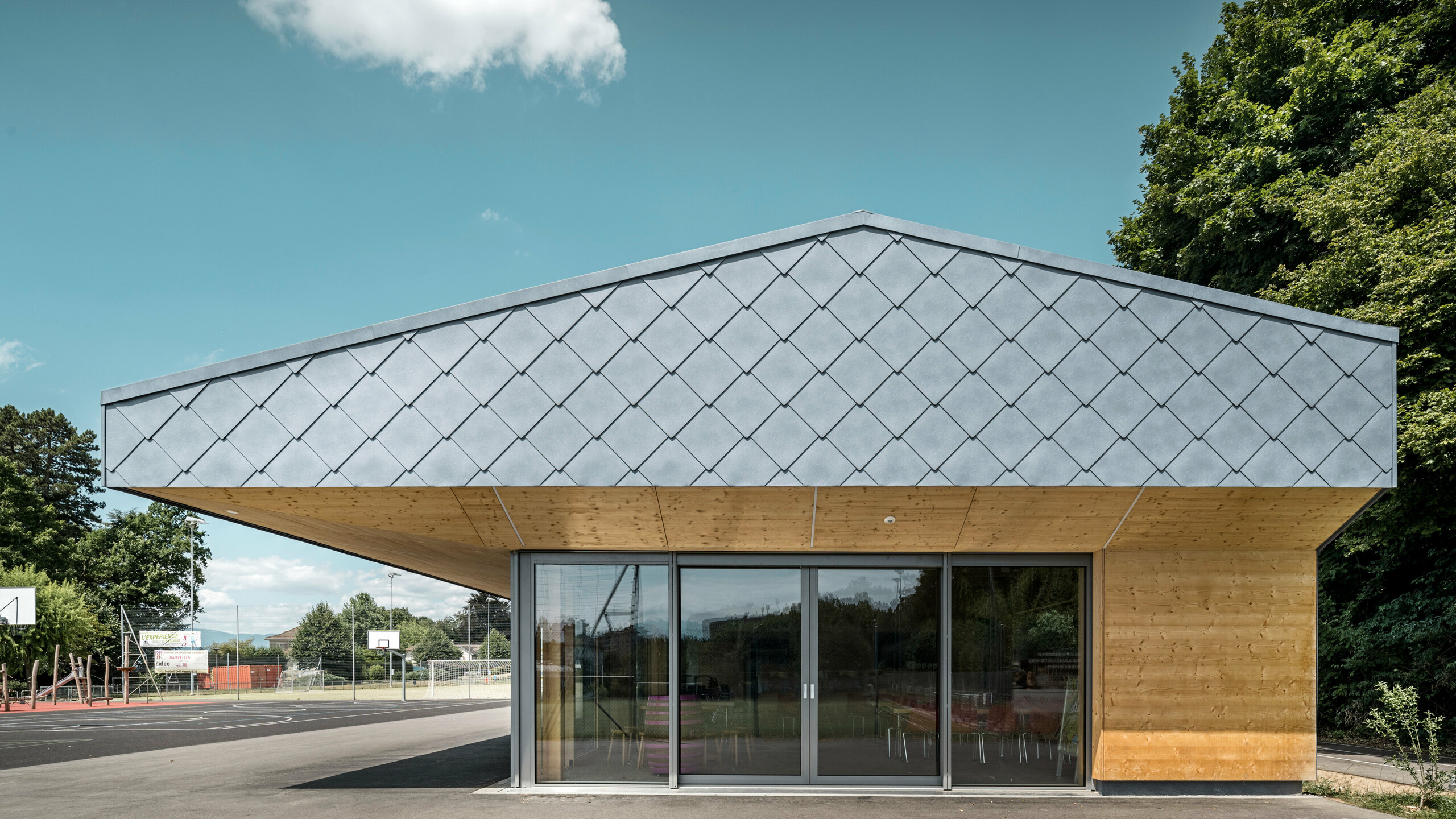 Seitenansicht der Sportanlage Fleur de Lys in Prilly, Schweiz, mit einem markanten Vordach, geschutzt durch PREFA Wandrauten 44x44 in P.10 Steingrau. Das Bauwerk zeichnet sich durch  die strukturierte Oberfläche der Rauten aus, die ein modernes und dynamisches Erscheinungsbild schaffen. Die sichtbare Holzverkleidung der überdachten Fassade ergänzt die kühle Eleganz des Aluminiums und unterstreicht die natürliche Schönheit des Gebäudes, während die Glasfronten für Transparenz und Offenheit stehen.