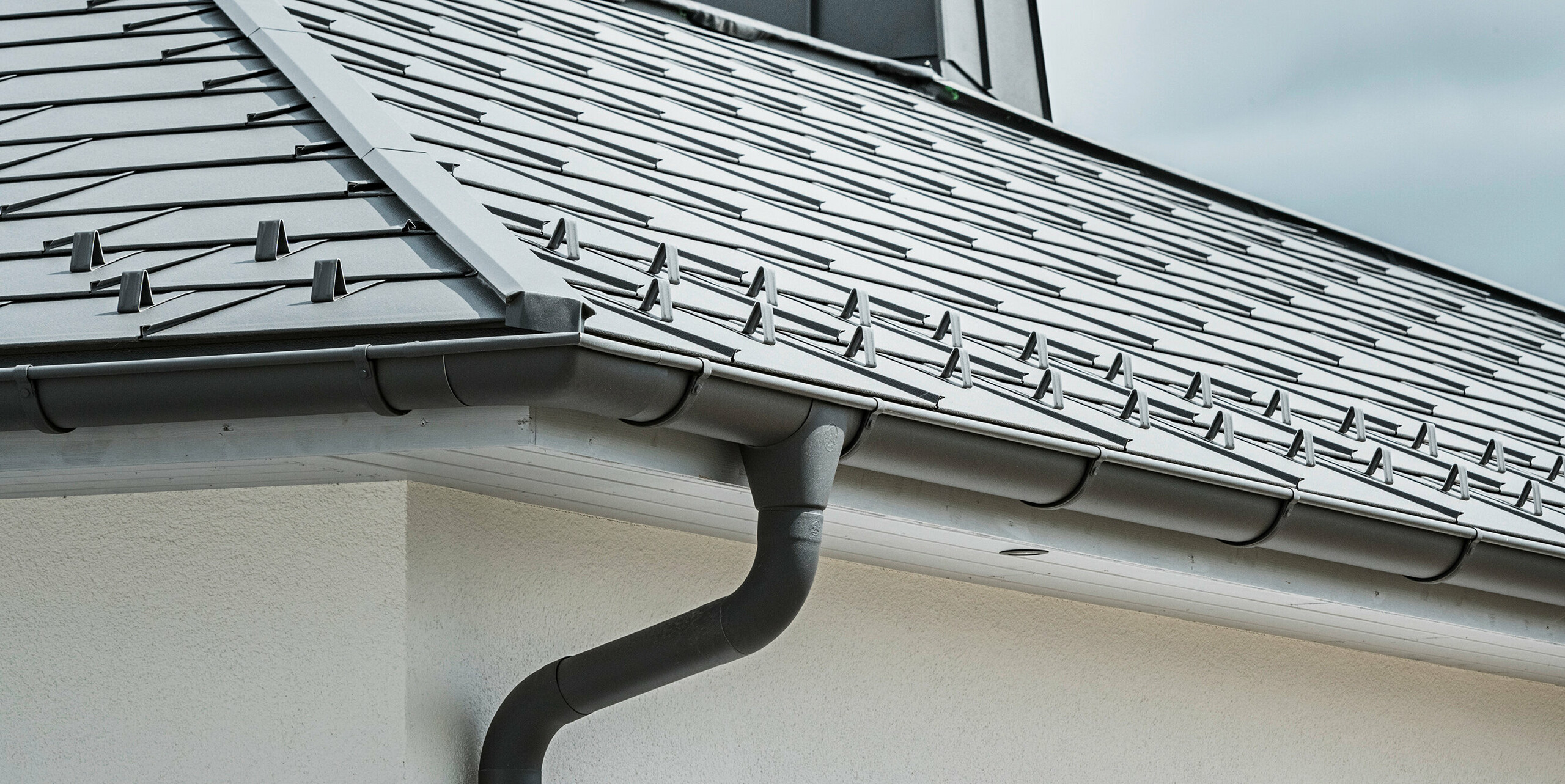 Nærbilde av takskjegget på en enebolig, utstyrt med det effektive PREFA takavløpssystemet i P.10 mørk grå. Bildet viser den detaljert monterte PREFA takplate R.16 og under den sømløst integrerte takrenne samt nedløpsrør, som sammen gir en pålitelig og estetisk tiltalende løsning for avledning av regnvann. De robuste aluminiumsproduktene og den nøye monteringen understreker kvaliteten og holdbarheten til PREFA komplett system.