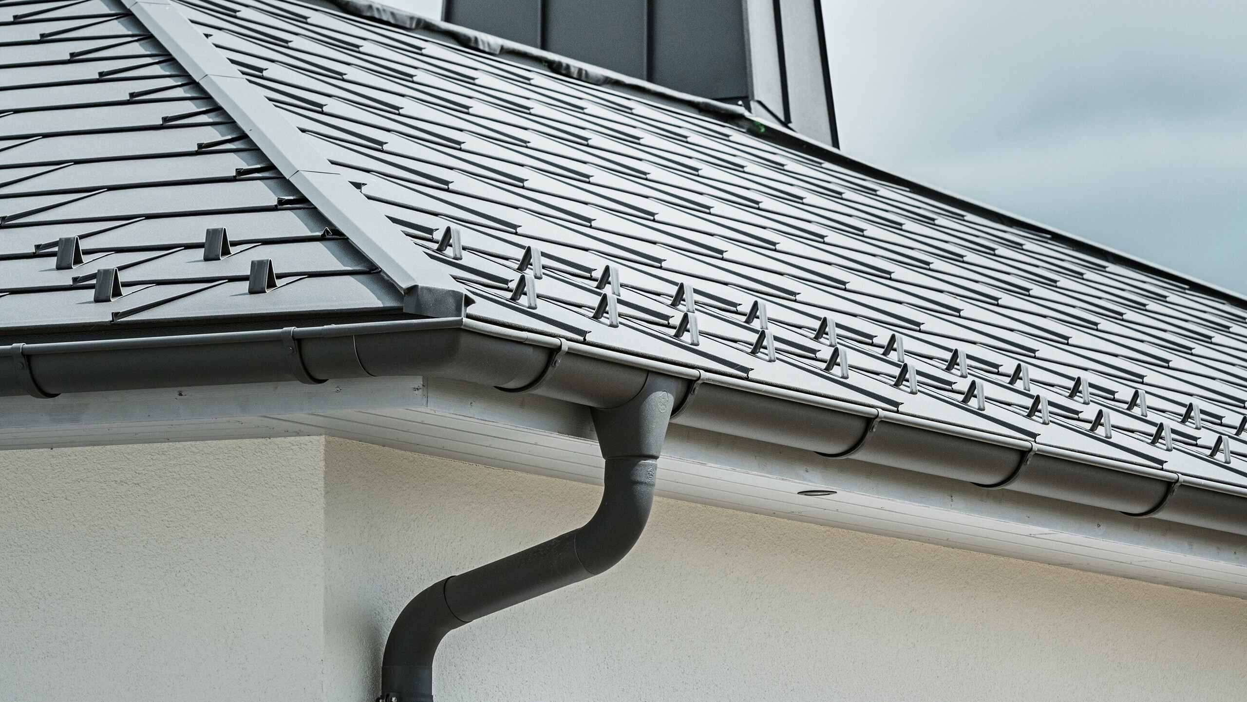 Nærbilde av takskjegget på en enebolig, utstyrt med det effektive PREFA takavløpssystemet i P.10 mørk grå. Bildet viser den detaljert monterte PREFA takplate R.16 og under den sømløst integrerte takrenne samt nedløpsrør, som sammen gir en pålitelig og estetisk tiltalende løsning for avledning av regnvann. De robuste aluminiumsproduktene og den nøye monteringen understreker kvaliteten og holdbarheten til PREFA komplett system.