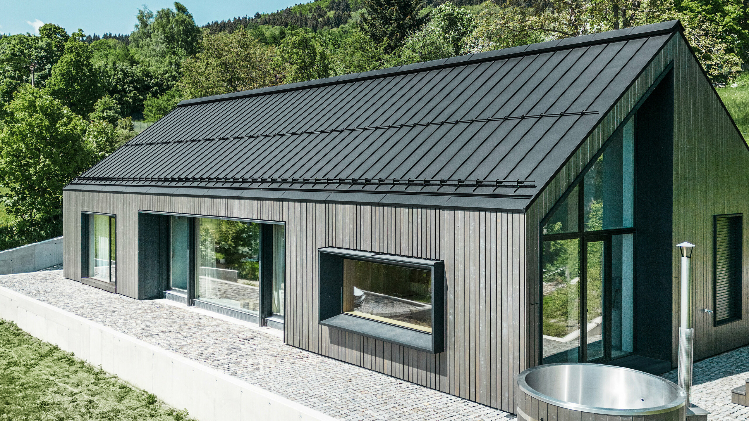 Ein modernes Holzmassivhaus in Kuncice (CZ) wurde mit großen Fensterflächen ausgestattet. Das dunkle Holzmassivhaus verfügt über ein Dachsystem aus PREFALZ in der Farbe P. 10 Schwarz. Im hinteren Außenbereich steht ein Whirlpool mit einer Holzverkleidung.