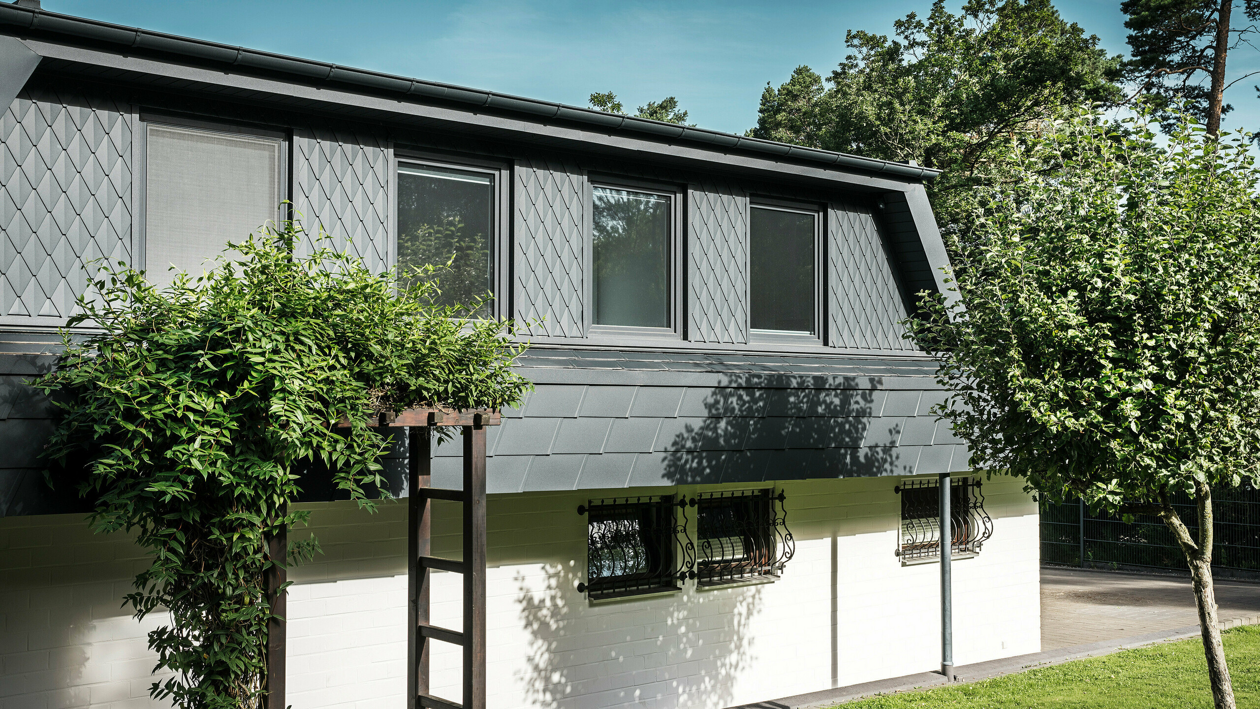 Seitenansicht eines Einfamilienhauses in Mechernich mit großzügigen Trapezgauben. Die Gauben sind mit kleinen PREFA Dachrauten in P.10 Anthrazit versehen, während das Dach mit den bewährten PREFA Dachschindeln DS.19 ebenfalls in P.10 Anthrazit eingedeckt ist. Die Aluminium-Dachrauten und -schindeln sorgen für eine wetterbeständige, ästhetische und nachhaltige Lösung für Dach und Fassade. Das traditionelle Einfamilienhaus ist von einem gepflegten Garten und zahlreichen Bäumen umgeben.