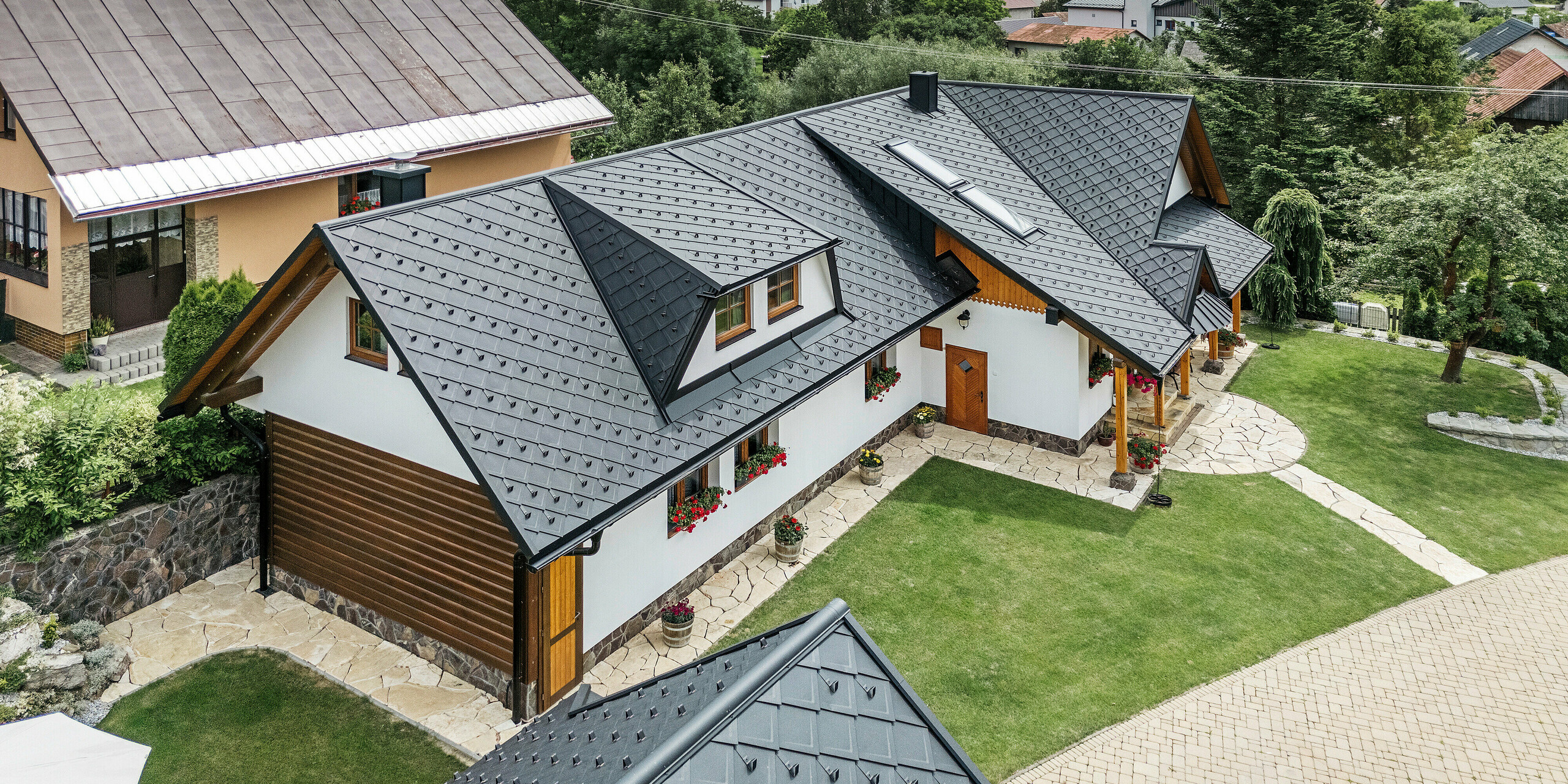 Luftaufnahme eines traditionellen Einfamilienhauses in Stara Bystrica, Slowakei, eingedeckt mit PREFA Dachrauten 29 × 29 in der Farbe P.10 Schwarz. Das Dach besticht durch seine elegante Schuppenoptik und die harmonische Integration von Gauben und Dachfenstern. Die umliegende grüne Landschaft, der gepflegte Garten und die gepflasterte Einfahrt unterstreichen das ansprechende Gesamtbild des Gebäudes.