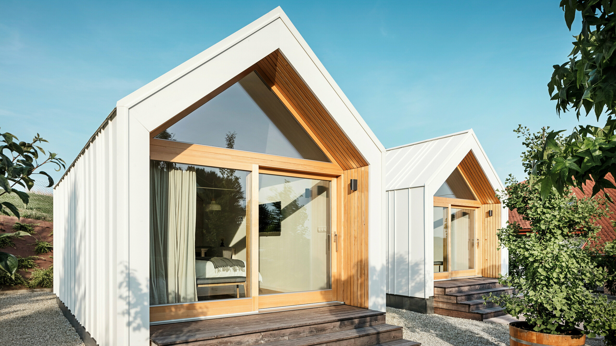 Frontansicht der beiden Tiny Häuser der Gästeunterkunft 'Pri Momi' in Kančevci, Slowenien, mit großen Glasfronten, gestaltet mit PREFALZ Dach- und Fassadenverkleidung in P.10 Reinweiß und naturbelassenem Holz. Die minimalistischen Mikrohäuser verfügen über breite Holzstufen, die zu den Eingängen führen, und bieten einen direkten Blick in die gemütlichen Innenräume. Die klare Linienführung und die Kombination aus Aluminium und Holz unterstreichen das moderne und einladende Design der Ferienunterkünfte.