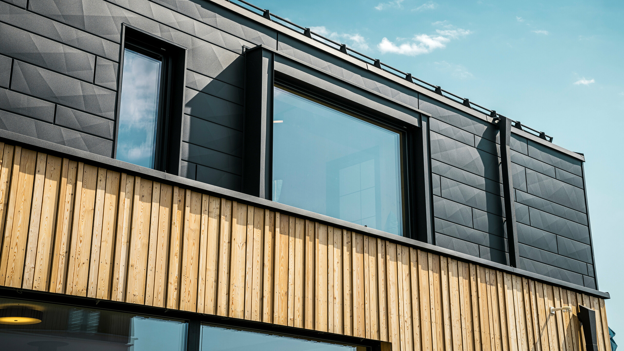 Nærbilde av øverste etasje i mønsterhuset Autark i prefabrikksenteret Blaue Lagune, Vösendorf (AT). Fasaden består av PREFA Siding.X aluminiumpaneler i P.10 svart, som skiller seg ut med sitt spesielle utseende med forskjellige kantninger. Under disse er det en stilfull trebekledning som danner en harmonisk kontrast til det mørke aluminiumet. Store vinduer slipper mye lys inn i bygningen og understreker det moderne designet.