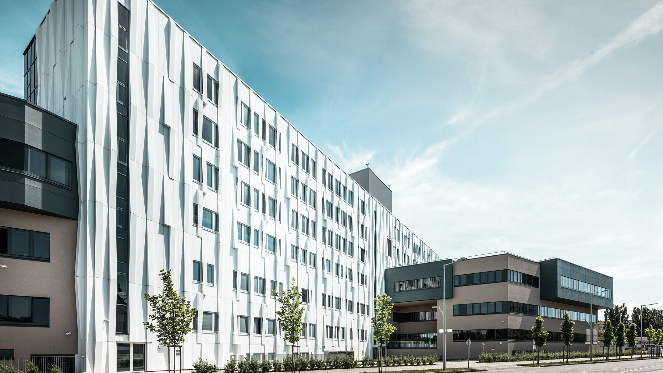 Universitetsklinikkens fasade sett fra siden. En vei med noen trær vises i forgrunnen.