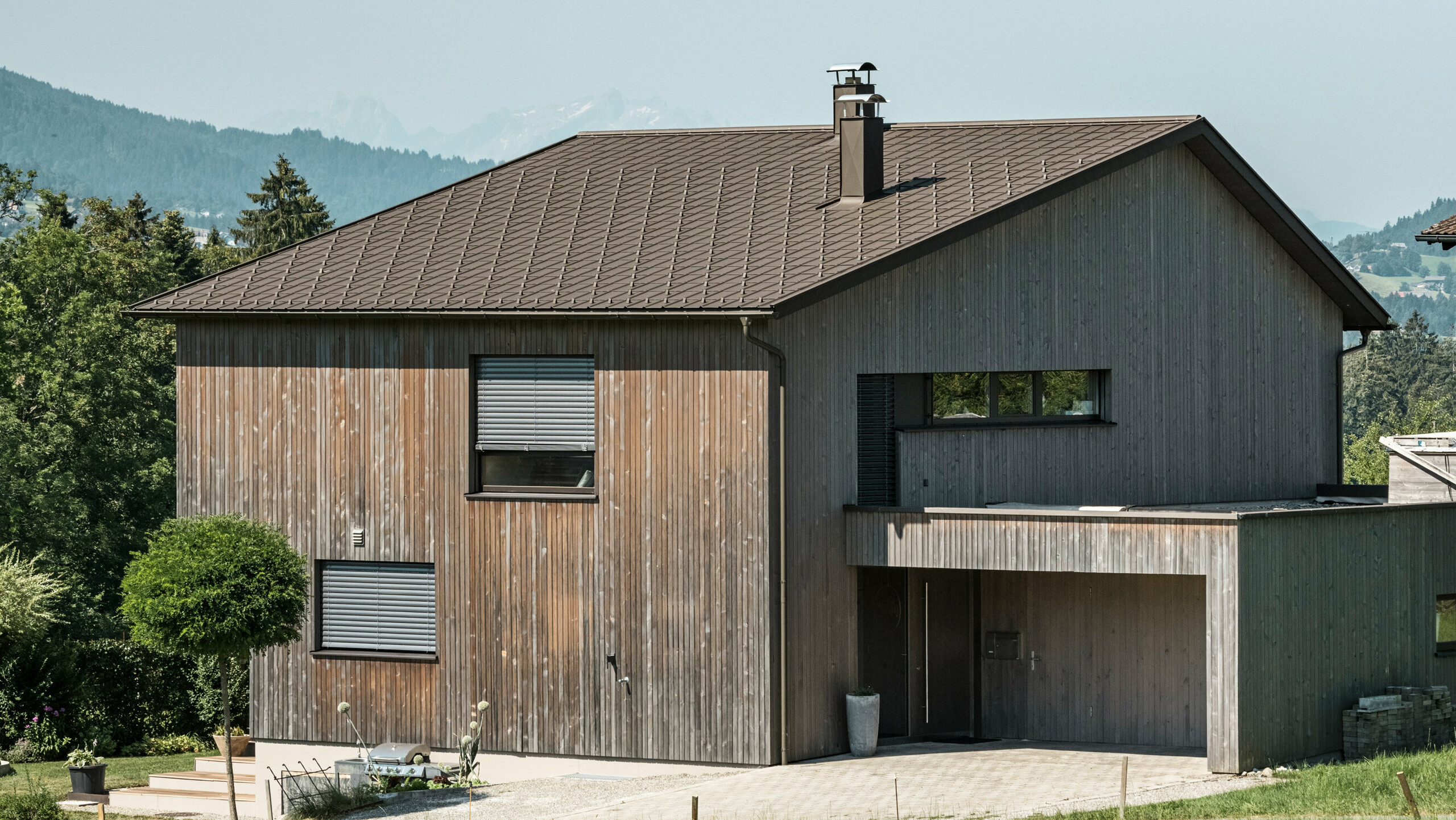Sidebilde av en moderne enebolig i Krumbach med en kombinasjon av en naturlig trefasade og et slitesterkt PREFA-tak i aluminium i fargen P.10 brun. Det harmoniske fargevalget viser den elegante integrasjonen av moderne design og tradisjonelle materialer, og PREFAs takplater gir en estetisk og funksjonell takdesign.