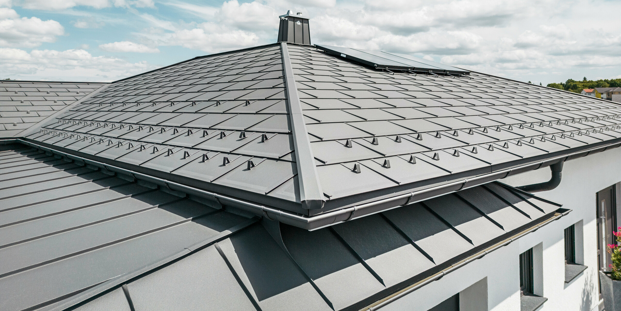 Detailansicht eines PREFA Aluminiumdachs auf einem Einfamilienhaus in Ilmenau, Thürigen. Das Dach wurde überwiegend mit PREFA Dachplatten R.16 in P.10 Dunkelgrau eingedeckt. Die kleinformatigen Aluminiumdachplatten wurden fachmännisch verlegt und verleihen dem Bungalow einen modernen Look. Das Bild zeigt die Präzision des PREFA Dachsystems, inklusive der Schneefangsysteme, die für zusätzliche Sicherheit sorgen. Die harmonische Integration der Hängerinnen und Ablaufrohre des PREFA Dachentwässerungssystems betont die ästhetische und funktionale Qualität des Gebäudes. Am linken Bildrand sind Stehfalzstreifen zu sehen, welche mit dunkelgrauem PREFALZ auf der direkt anschließenden Garage montiert wurden.