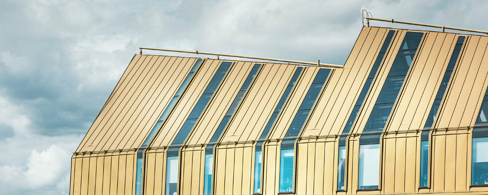 Moderner, mehrgeschossiger Bürobau mit charakteristischer goldfarbener Stehfalz‑Metalldach‑ und Fassadenverkleidung, vertikal gegliederte Fensterflächen und großflächige Dachflächenfenster, klare Linienführung und zeitgenössische Materialität vor bewölktem Himmel – repräsentativ für ein funktionales, anspruchsvolles Bürogebäude mit präziser Aluminiumverarbeitung und starker architektonischer Präsenz.