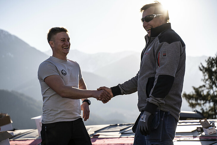 Auf einem geneigten Hausdach stehen ein PREFA-Trainer und ein Spengler, die sich die Hand geben. Beide tragen professionelle Arbeitskleidung und Sicherheitsausrüstung. Im Hintergrund sind eine tiefstehende Sonne und Verpackungsschachteln von PREFA-Aluminiumprodukten zu sehen. Diese Szene symbolisiert eine erfolgreiche Zusammenarbeit und einen fachlichen Austausch mit Handschlagqualität. Der PREFA-Trainer unterstützt den Spengler mit seinem Know-how zu den Themen Verarbeitung, Technik und Produktsysteme. Das Bild steht für die enge Betreuung von Handwerkern durch PREFA – von der Schulung bis zur professionellen Umsetzung auf der Baustelle.