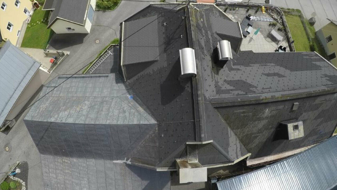 Fugleperspektiv over et gammelt tak med mange detaljer – søkk, kvister og skorstein. Det gamle takbelegget har blitt utbedret flere ganger og er flekkete.