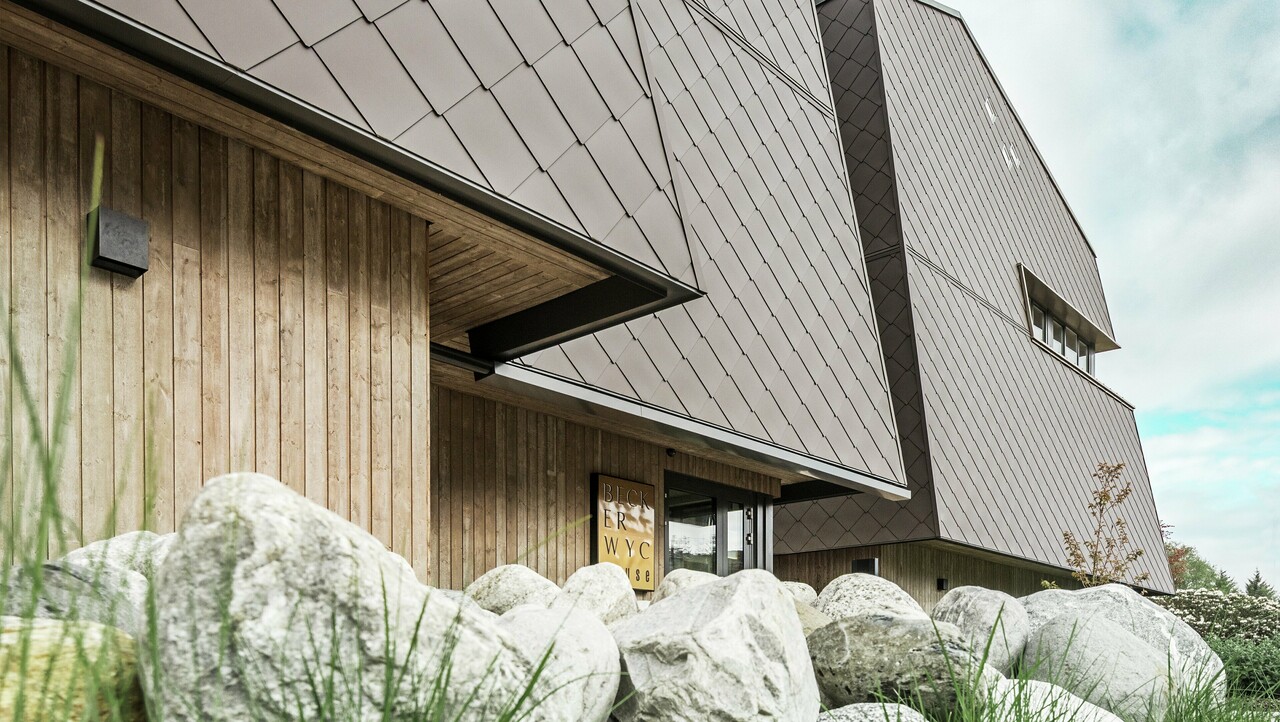 Sidebilde av et hotell, der fasaden i første etasje er kledd vertikalt med mørkfarget treverk. Over dette er det et fasadefremspring, og herfra er fasaden kledd med aluminiumspaneler i valnøttbrun.