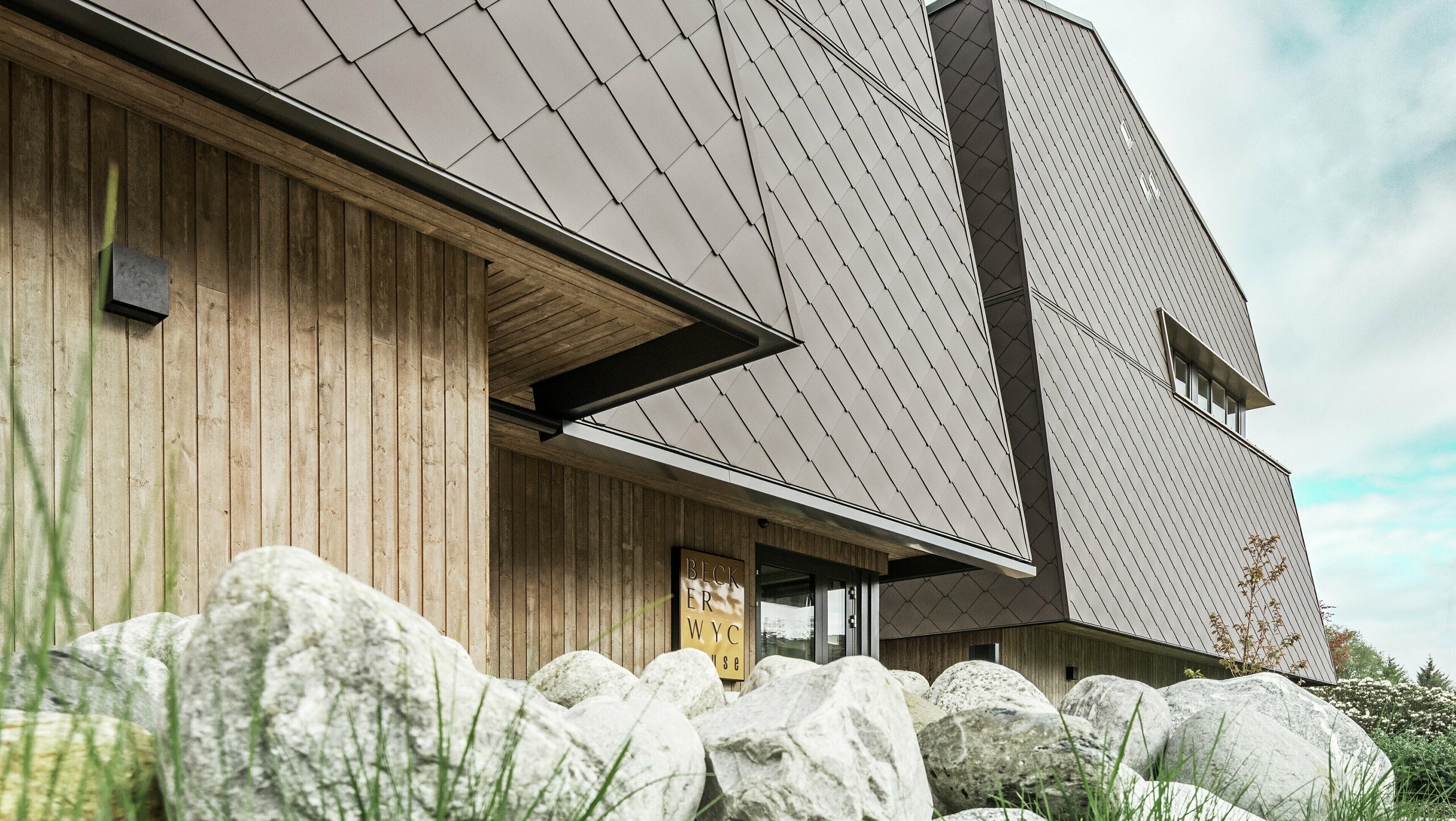 Sidebilde av et hotell, der fasaden i første etasje er kledd vertikalt med mørkfarget treverk. Over dette er det et fasadefremspring, og herfra er fasaden kledd med aluminiumspaneler i valnøttbrun.