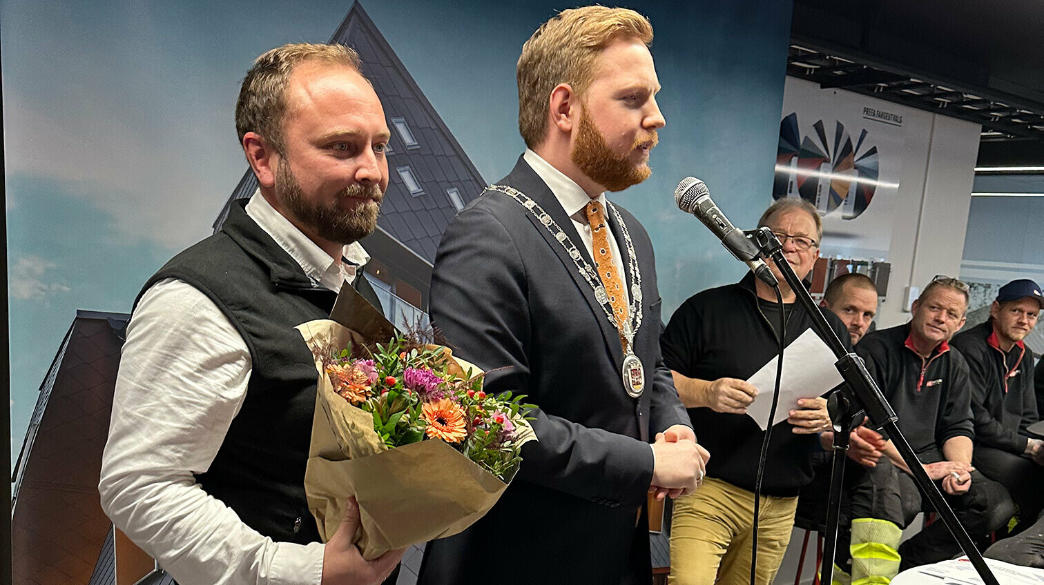 Der Bürgermeister von Skien (rechts) hält eine Rede während der Eröffnung der PREFA Academy in Skien. Neben ihm steht ein PREFA-Vertreter, der einen Blumenstrauß hält. Im Hintergrund hören weitere Teilnehmer der Veranstaltung aufmerksam zu.