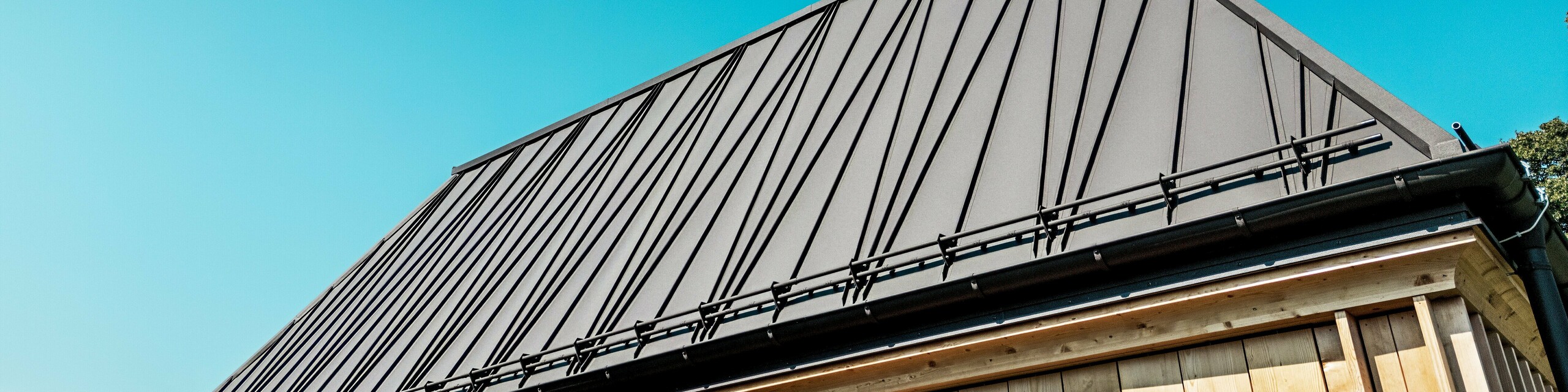 Nahaufnahme des außergewöhnlichen PREFA Stehfalzdachs des „Badehauses“ beim Hotel Hirschen in Schwarzenberg. Das Dach in der Farbe P.10 Nussbraun besticht durch seine markanten, teilweise diagonal verlaufenden Stehfalze, die dem Gebäude eine dynamische, moderne Ausstrahlung verleihen. Die klare Linienführung und die 60-Grad-Dachneigung erzeugen eine kraftvolle geometrische Wirkung. Unter dem Dach ist die Fassade mit vertikal angeordneten Holzlamellen sichtbar, die traditionelle Bauweise neu interpretieren. Der wolkenlose Himmel und das direkte Sonnenlicht betonen die hochwertige Oberfläche des PREFALZ Materials sowie das präzise Spenglerhandwerk. Das Bild fängt die Verbindung von Innovation, Handwerk und regionalem Stil eindrucksvoll ein.