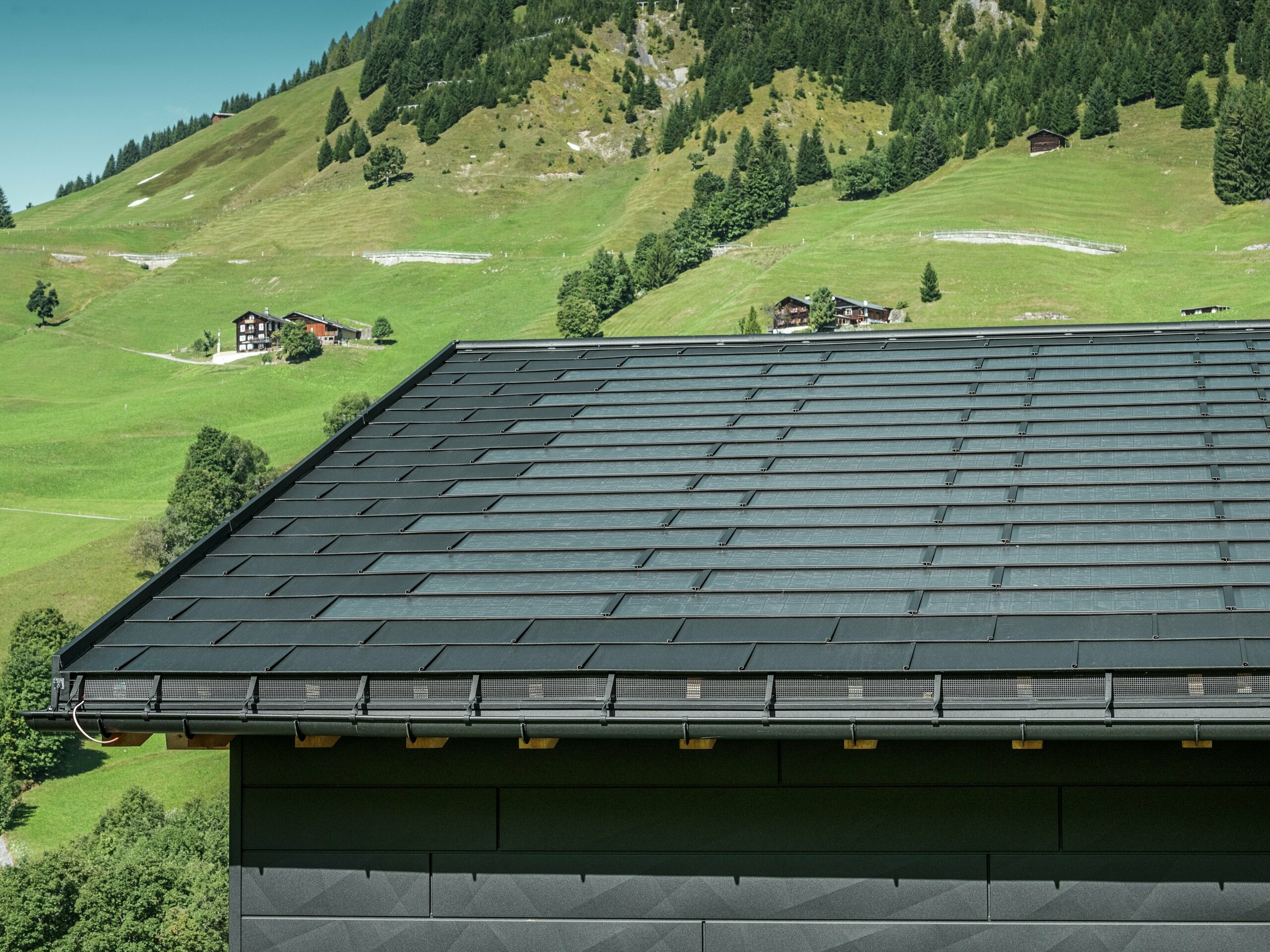 Nærbilde av takskjegget på en moderne enebolig i de alpine omgivelsene i Ascharina i Sveits. Taket er dekket med den innovative PREFA solcelle takplate og den tradisjonelle takplate R.16, begge i P.10 svart. PREFA takrenne og PREFA nedløpsrør sørger for pålitelig takdrenering. Nederst på bildet ser du de karakteristiske PREFA Siding.X fasadepaneler. De klare linjene i aluminiumsproduktene understreker det moderne designet midt i et pittoresk fjellandskap.