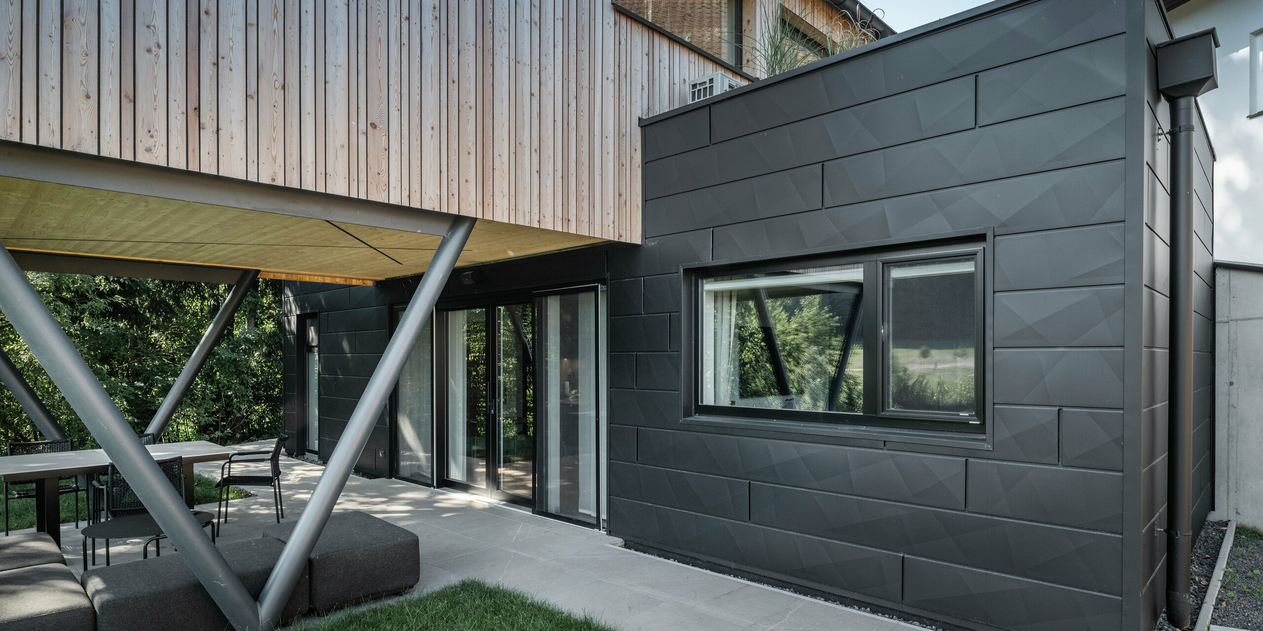 Arkitektonisk høydepunkt: «Woodstock Chalet 5» i St. Martin am Innkreis, Øvre Østerrike. Det moderne bryllupsstedet fremheves ved bruk av antrasittfargede PREFA Siding.X fasadepaneler. Aluminiumet danner en sterk kontrast til de lyse trepanelene og de metalliske støttestrukturene. De mørke vindusrammene passer harmonisk inn i fasadedesignet, noe som gir bygningen et elegant utseende. Dette bildet viser den estetiske balansen mellom naturlig tretekstur og den industrielle sjarmen til aluminiumsfasaden, som står for designet av moderne bygninger.