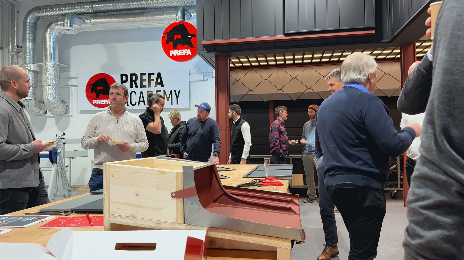 Teilnehmer der PREFA Academy Eröffnung in Skien, die sich in der modernen Trainingsumgebung von PREFA austauschen. Im Vordergrund ist ein PREFA-Dachsystem zu sehen, während im Hintergrund das PREFA Academy-Logo an der Wand zu sehen ist. 