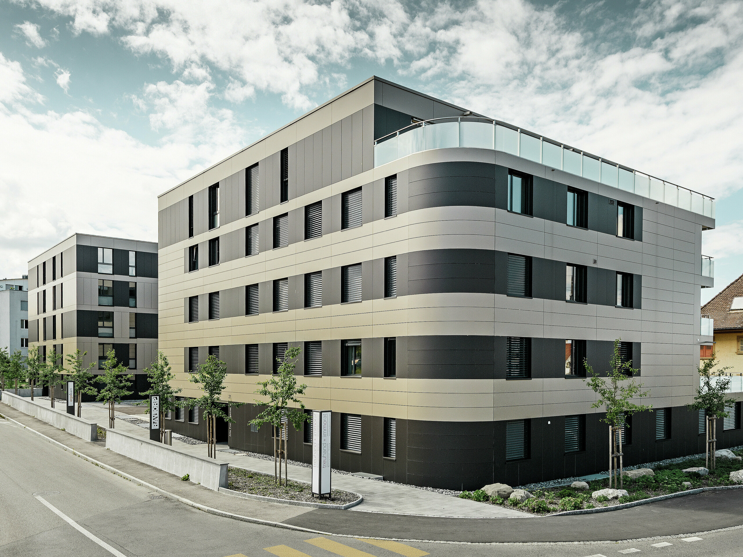 In der kubischen Wohnanlage von Murten (CH) sorgen die PREFA Aluminium Verbundplatten in Bronze und Schwarzgrau für eine optische Aufwertung und gleichzeitig für eine robuste und langlebige Fassade. Die Kombination dieser Farbtöne bietet ästhetische Akzente und schützt das Gebäude langfristig vor Wettereinflüssen. An der Gebäudeecke zur Straße hin ist eine Rundung zu erkennen, was das Rollbiegen der PREFABOND Aluminium Verbundplatten demonstrieren soll.