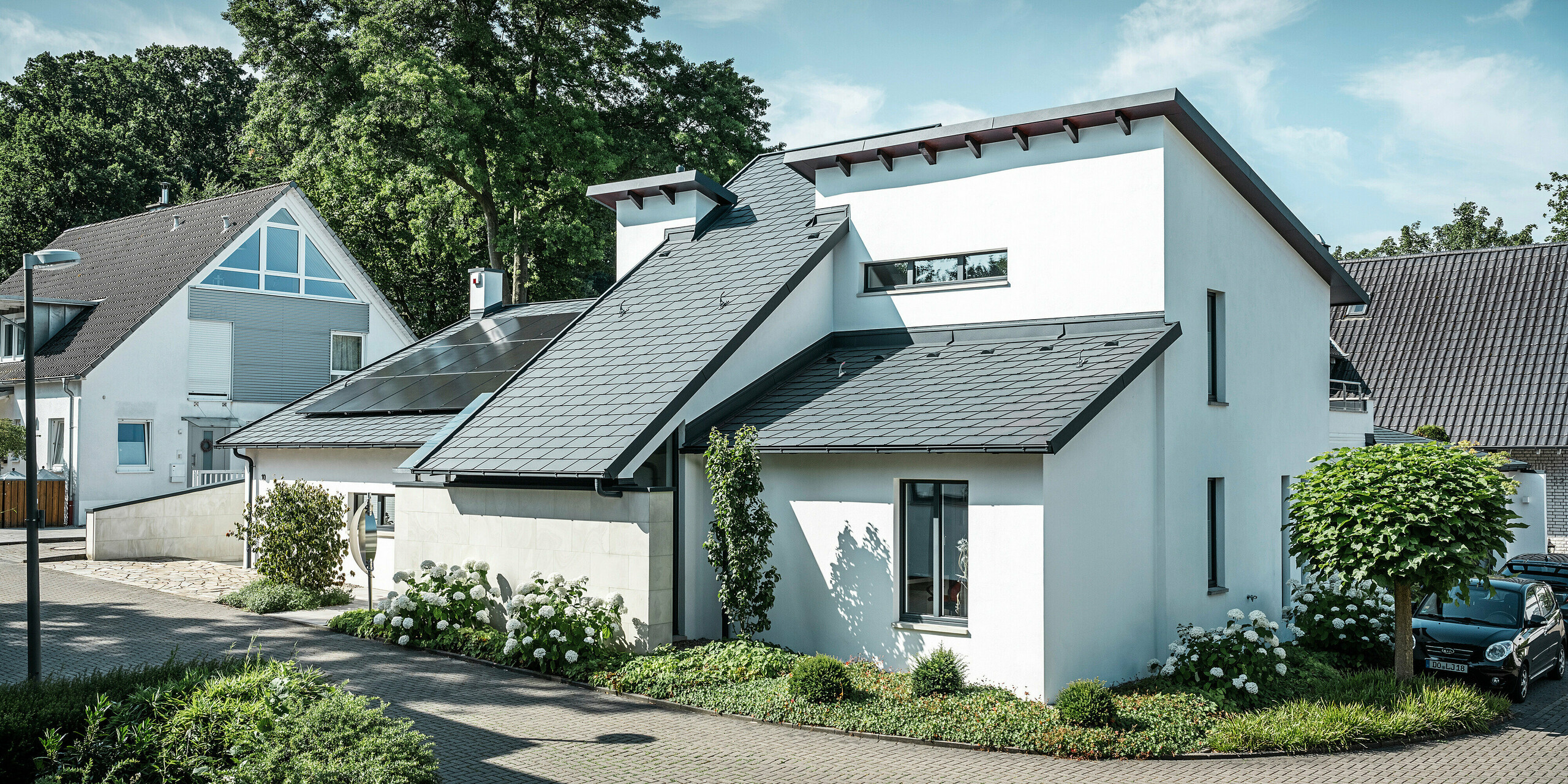 Modernes Einfamilienhaus mit weißer Putzfassade und dunklem PREFA Schrägdach aus Aluminium. Auf dem Dach sind integrierte Photovoltaikmodule und eine Aluminium-Dachentwässerung in Anthrazit zu sehen. Das Gebäude liegt in einer gepflegten Wohnsiedlung mit bepflanzten Vorgärten, gepflasterter Zufahrt und umliegenden Einfamilienhäusern.