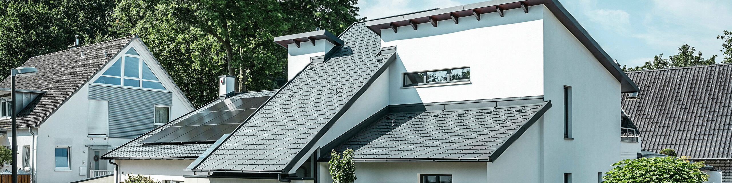Modernes Einfamilienhaus mit weißer Putzfassade und dunklem PREFA Schrägdach aus Aluminium. Auf dem Dach sind integrierte Photovoltaikmodule und eine Aluminium-Dachentwässerung in Anthrazit zu sehen. Das Gebäude liegt in einer gepflegten Wohnsiedlung mit bepflanzten Vorgärten, gepflasterter Zufahrt und umliegenden Einfamilienhäusern.