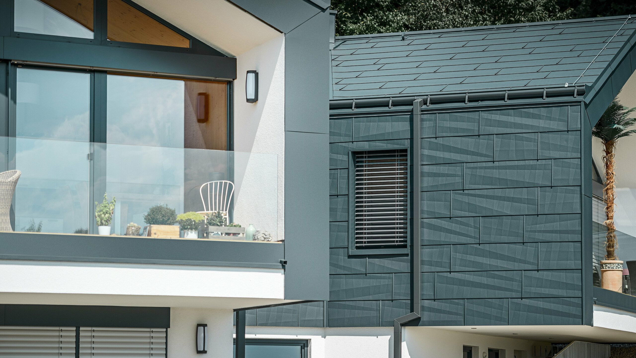 Bildet viser et moderne tomannsbolig med en stilfull fasade i aluminium. Fasaden er holdt i en mørk antrasittfarge og har en strukturert overflate takket være PREFA tak- og fasadepanel FX.12. På taket er det montert en fotrenne og et firkantrør som nedløpsrør fra PREFA. På øverste etasje er det en balkong med glassrekkverk, mens vinduene i underetasjen har lamellgardiner. Arkitekturen er klar og minimalistisk, og i bakgrunnen ser man trær som skaper en naturlig atmosfære.