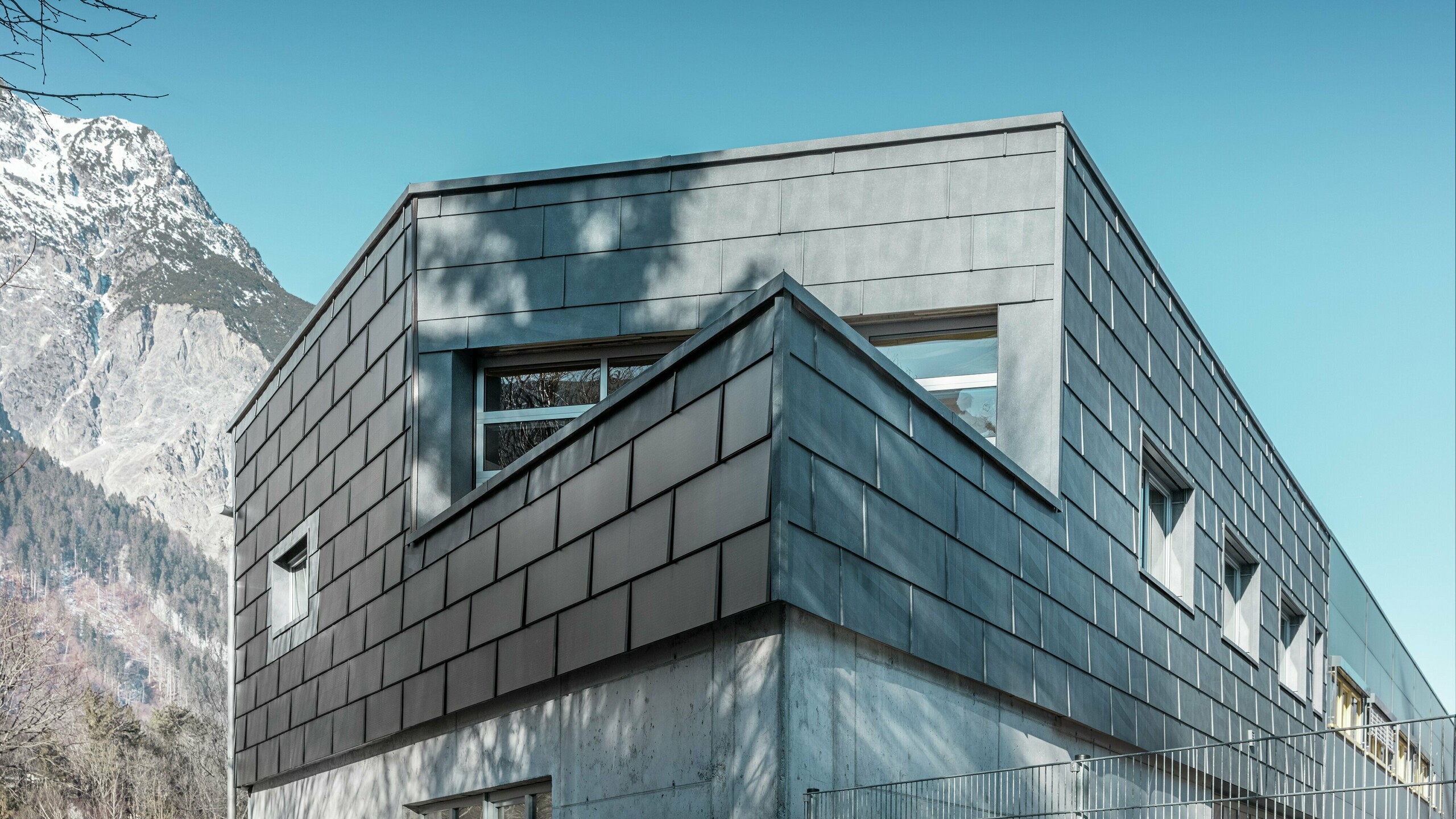 Das Firmengebäude der Spenglerei RÜMA in Vorarlberg beeindruckt mit seiner modernen Fassade aus PREFA Solardachplatten und Sichtbeton. Umgeben von Bergen und Bäumen, deren Schatten sich auf der Wand abzeichnen, steht es unter einem klaren, blauen Himmel. Die spitze Gebäudeecke und die PREFA Fassadenpaneele FX.12 betonen das markante Erscheinungsbild zusätzlich.