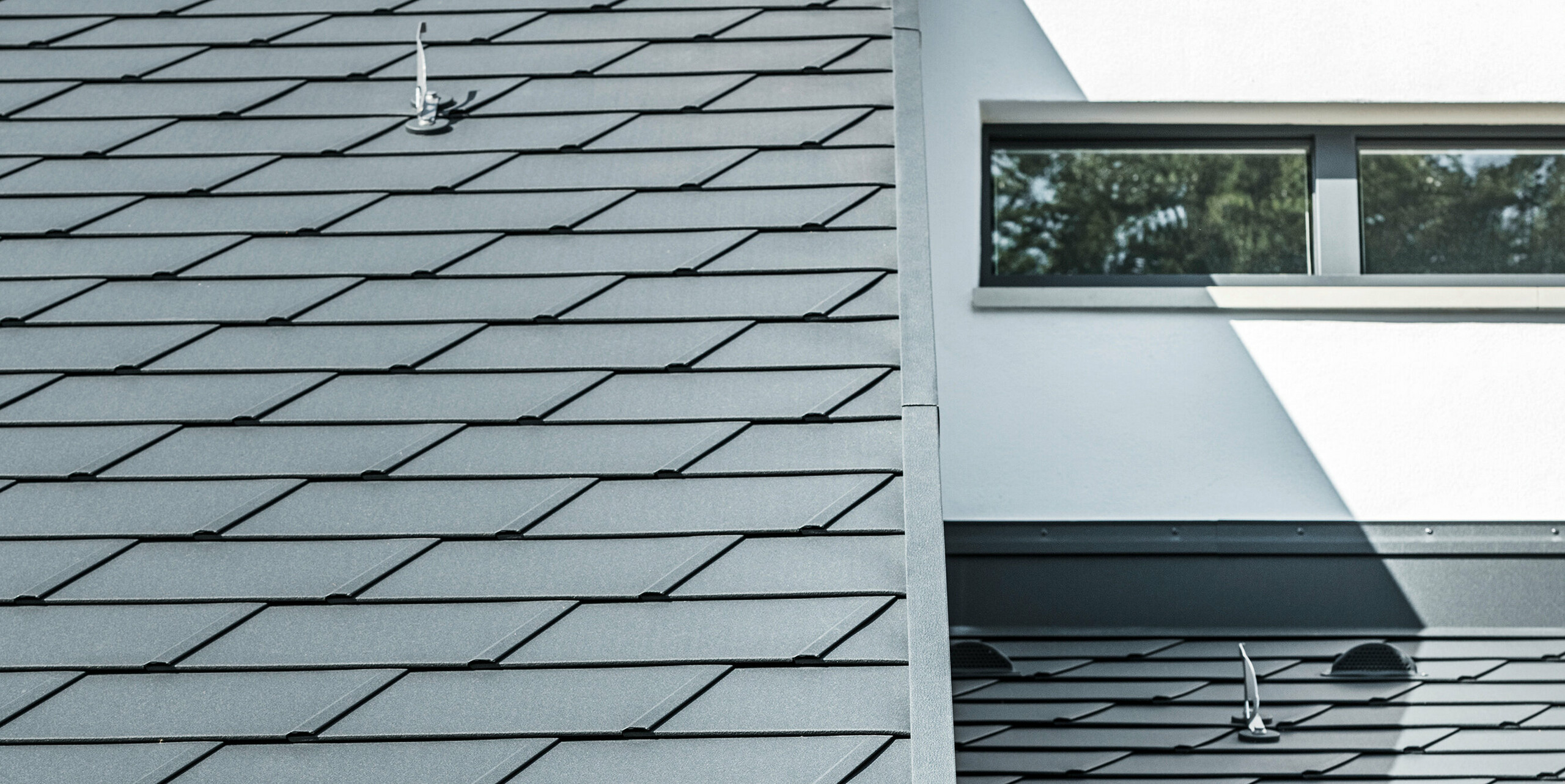 Detailaufnahme der unterschiedlichen Ebenen am Dach eines Einfamilienhauses in Dortmund. Zu sehen sind die PREFA Dachschindel DS.19 und das PREFA Dachentwässerungssystem in der Farbe P.10 Anthrazit sowie die PREFA Sicherheitsdachhaken.