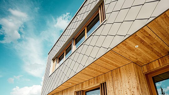 Die Nahaufnahme der Kindertagesstätte in Alzenau zeigt die präzise Ausführung und hochwertige Materialkombination aus bronzefarbenen PREFA Wandrauten 44 × 44 und naturbelassenem Holz. Die diagonale Struktur der Aluminiumfassade bildet ein markantes Schuppenmuster, das sich harmonisch mit den vertikalen Holzlamellen an den Fenstern und der fein ausgearbeiteten Holzuntersicht verbindet. Die Aufnahme aus der Froschperspektive betont die exakte Linienführung und handwerkliche Qualität der vorgehängten hinterlüfteten Fassade. Diese Detailansicht unterstreicht die gestalterische Stärke und Langlebigkeit von PREFA Produkten – ideal für nachhaltige Architekturprojekte, die Ästhetik, Funktionalität und Ökologie auf höchstem Niveau vereinen.